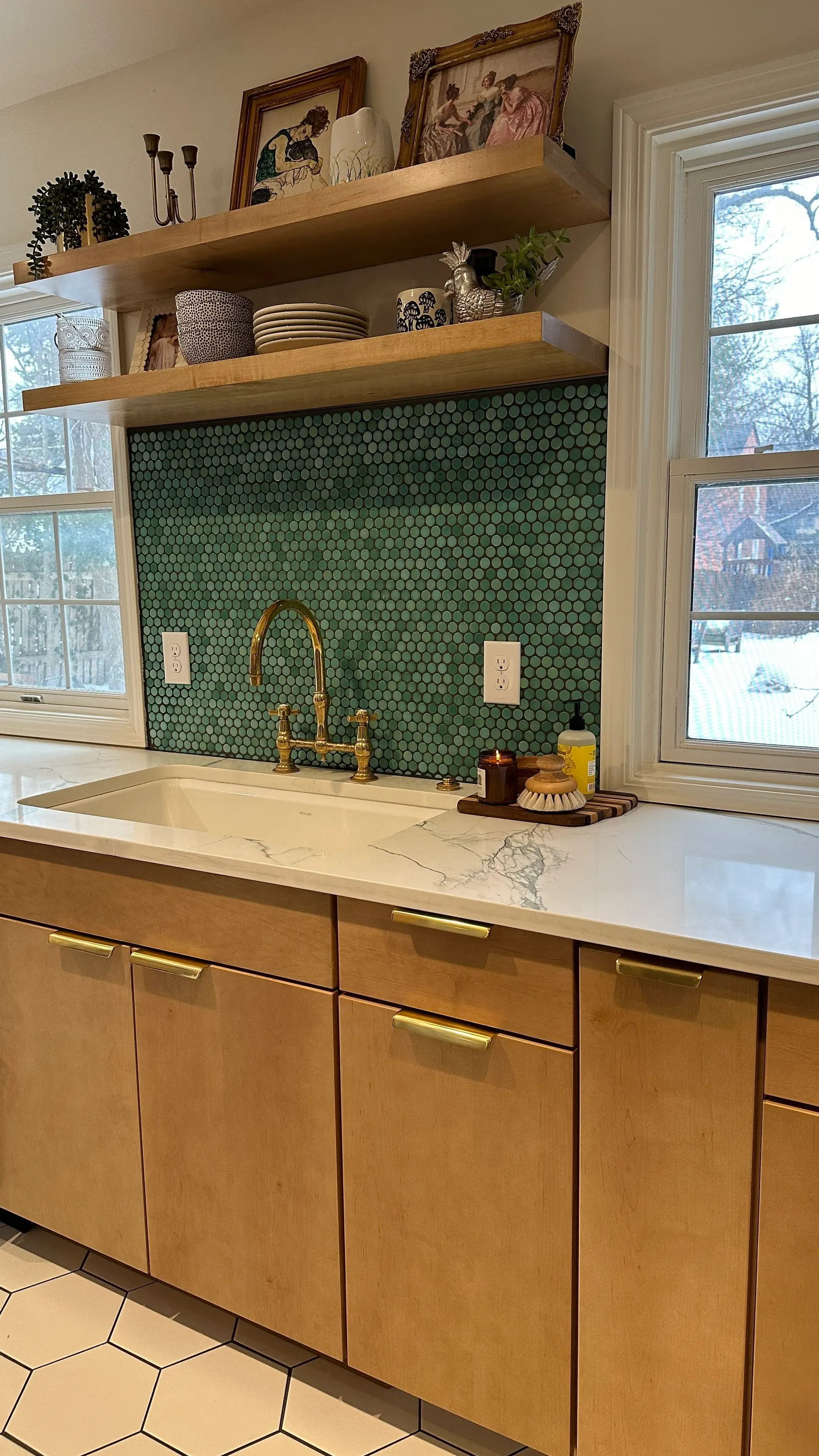 Kitchen sink with gold faucet, green tile backsplash, wooden cabinets, white marble countertop, open wooden shelves with dishes and decorative items, and two windows showing an outdoor snowy scene.