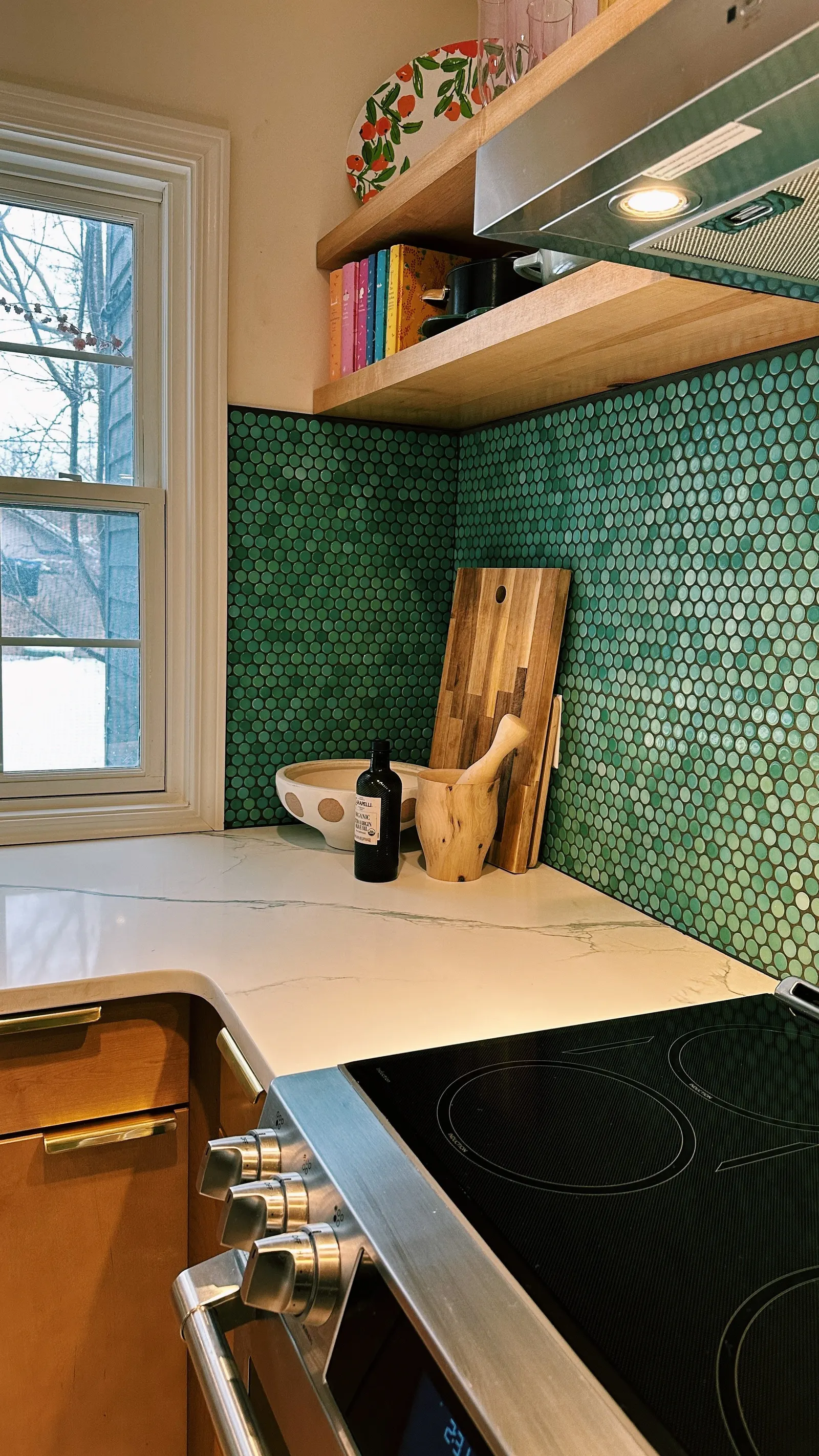 Kitchen corner with green circular tile backsplash, wooden shelves holding colorful books and cookware, a window with a snowy view, and a white marble countertop with kitchen utensils and a black electric stove.
