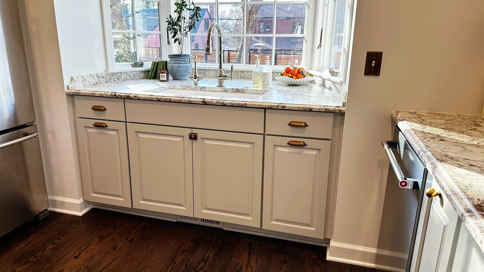 Kitchen sink with windowed view, countertop with potted plant, soap bottles, a fruit bowl, and decorated cabinets.