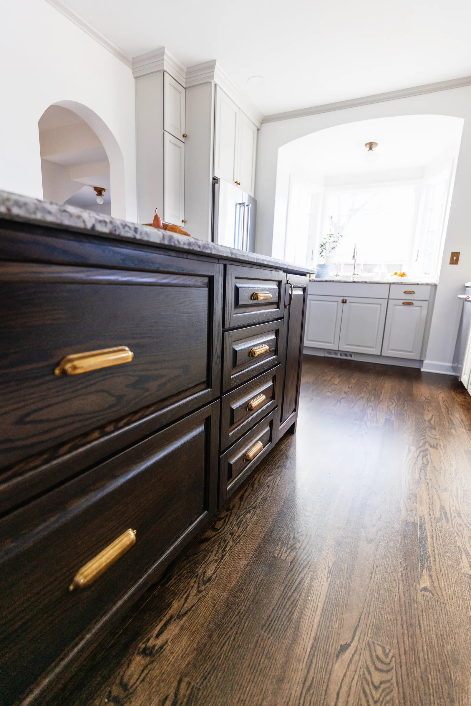 View of a kitchen with dark wooden cabinets with gold handles, white upper cabinets, a granite countertop, and bright natural light coming from a large window.