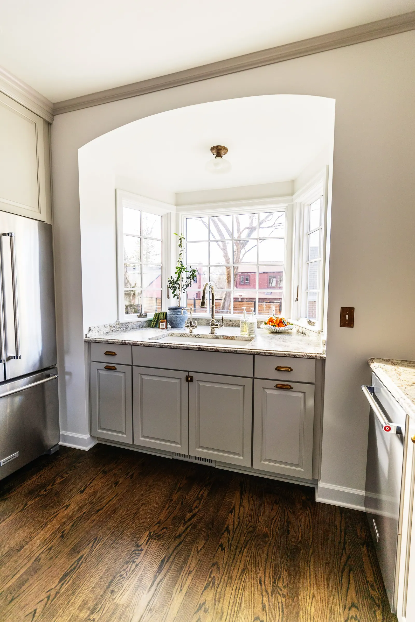 Bright kitchen with white cabinetry beneath a large bay window; wooden floors, stainless steel refrigerator, and granite countertops with a sink and potted plant.