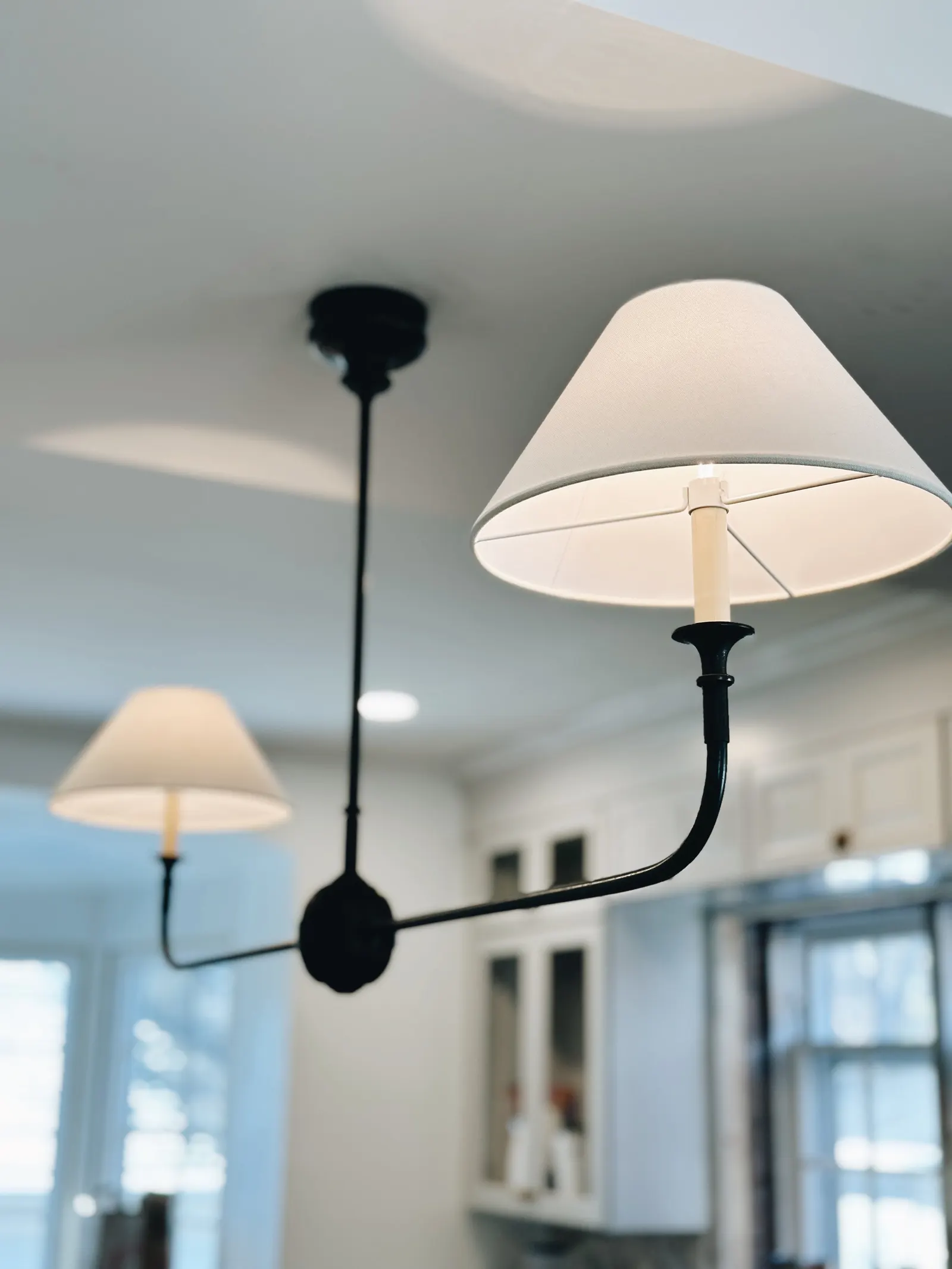 A close-up of a white ceiling lamp with two lampshades, featuring a black metal arm extending from a central mounting point in the ceiling, with a background of a white room with cabinets and windows.