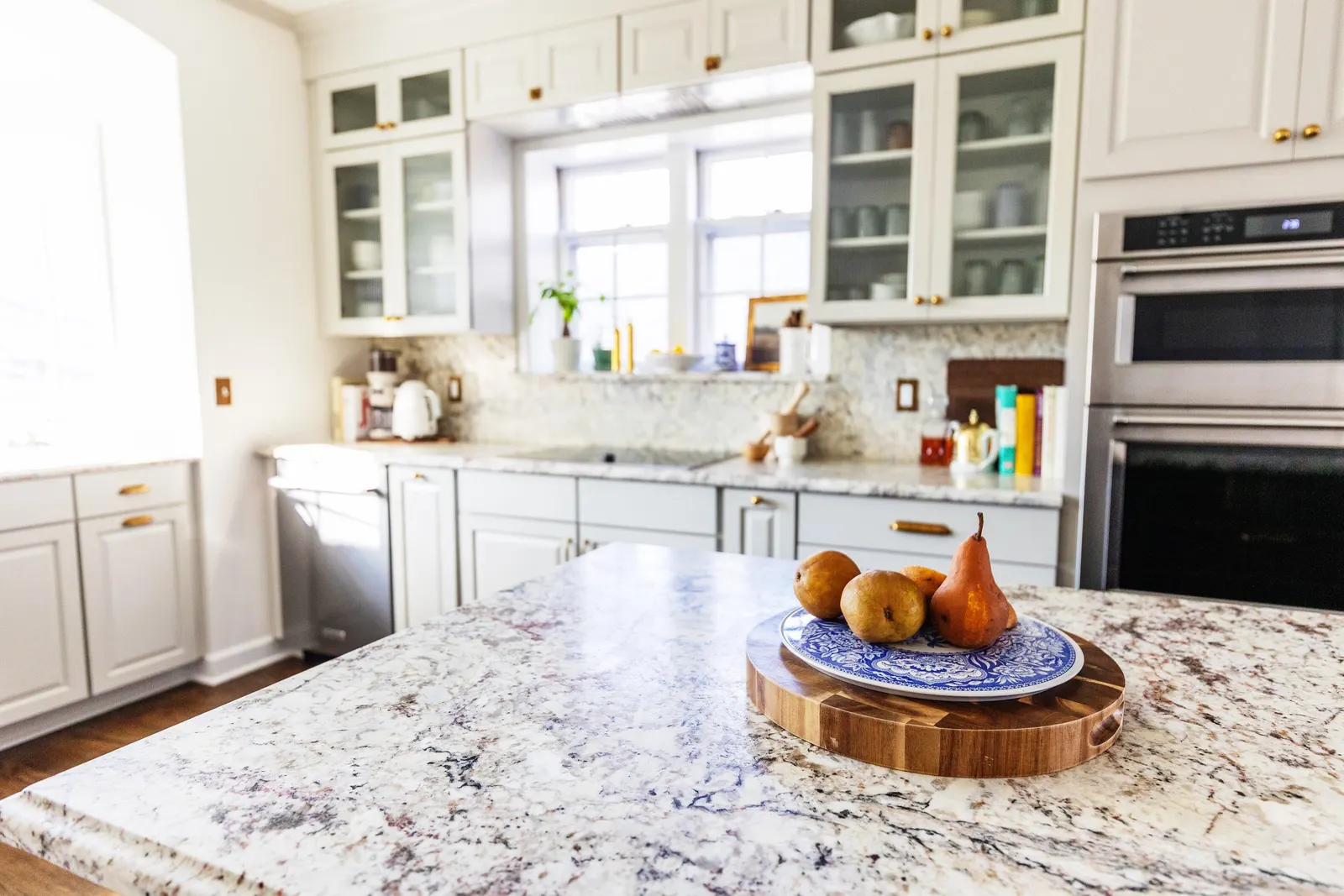 Kitchen with white cabinets, granite countertops, and a window. The kitchen island has a bowl of pears and apples on a blue-and-white plate on a wooden tray.