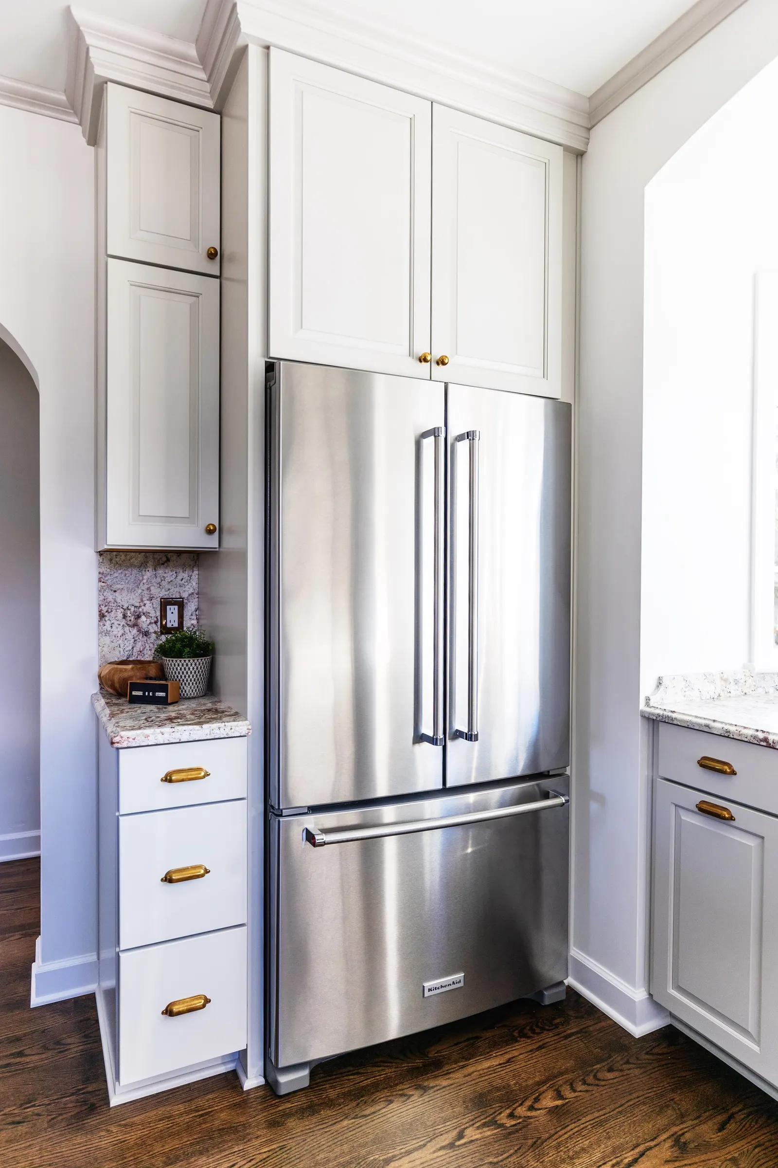 Stainless steel KitchenAid refrigerator surrounded by white cabinets with gold handles and granite countertops, in a kitchen with hardwood flooring.