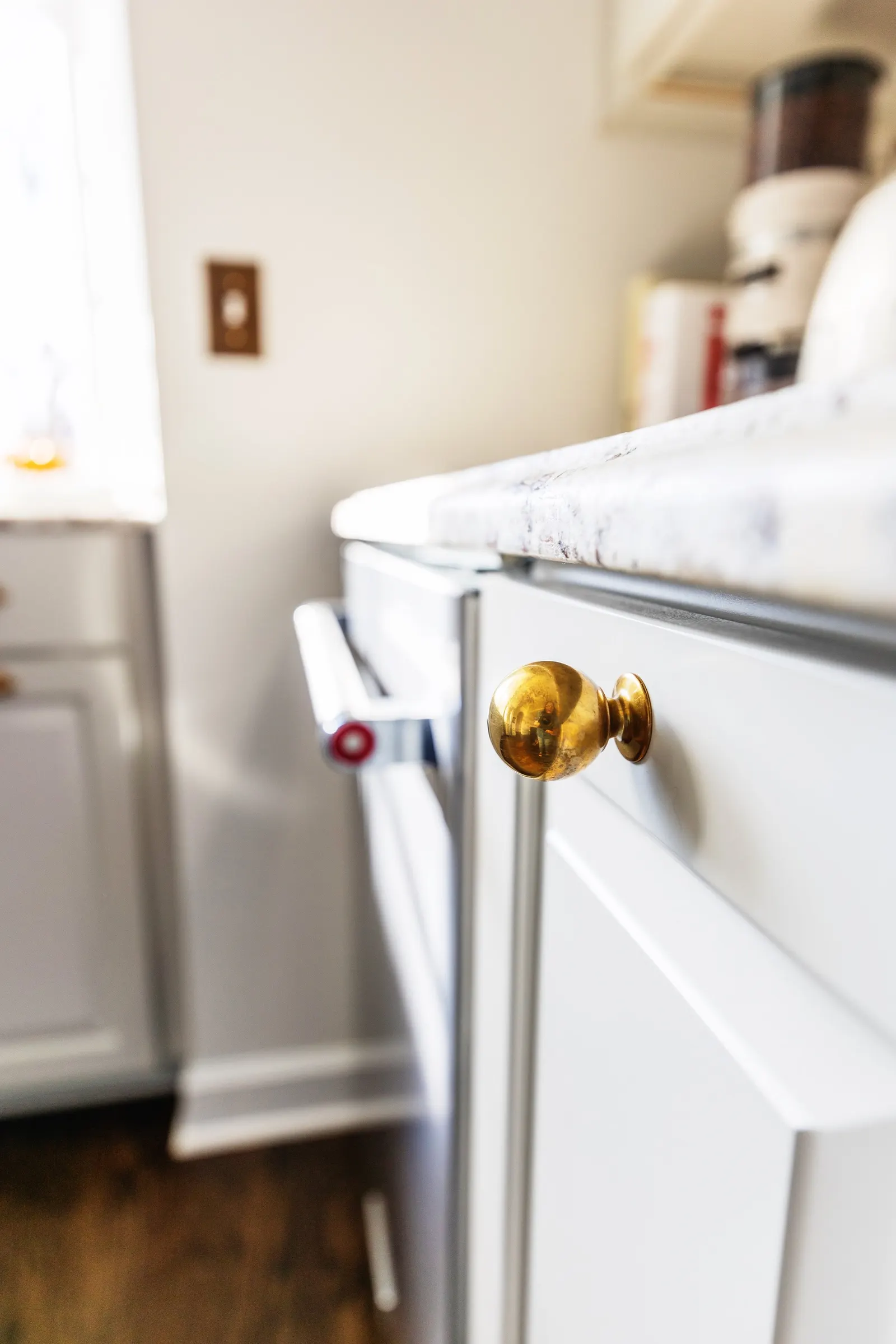 Close-up of a white kitchen cabinet with a shiny gold doorknob and a countertop above. Part of a stove is visible to the left, and a window with sunlight is in the background.