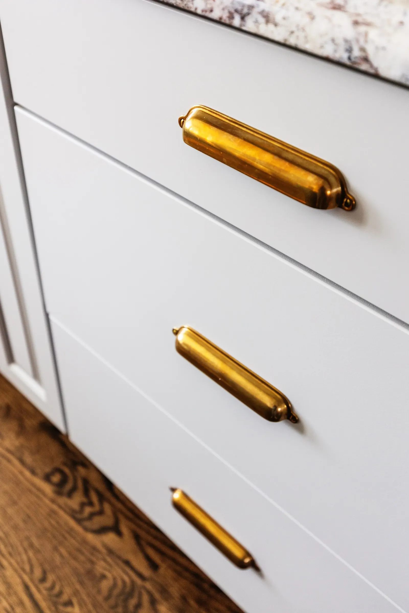 Close-up of a white dresser with two drawers, featuring gold handles. Part of a granite countertop and wooden floor are visible.