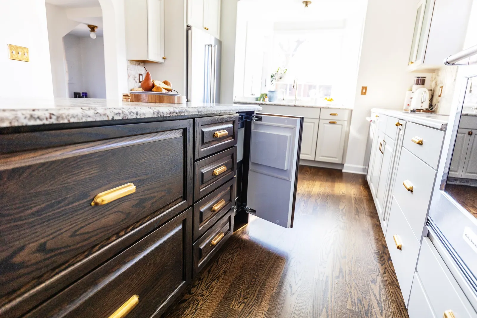 Kitchen with black and white cabinets, gold handles, granite countertops, and a wooden floor, with sunlight coming through a window.
