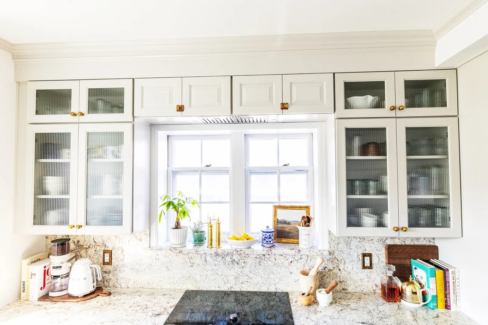 White kitchen with granite countertops, white cabinets with glass doors, and a window sill with plants, books, and decorative items.