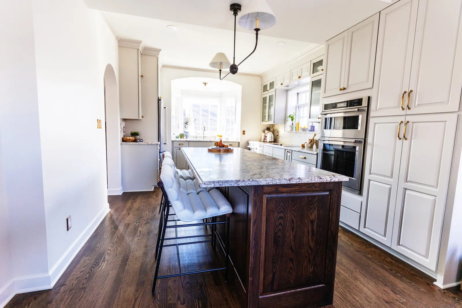 Bright kitchen with white cabinets, a granite island with dark wood base, and a double oven, illuminated by natural light from a bay window.