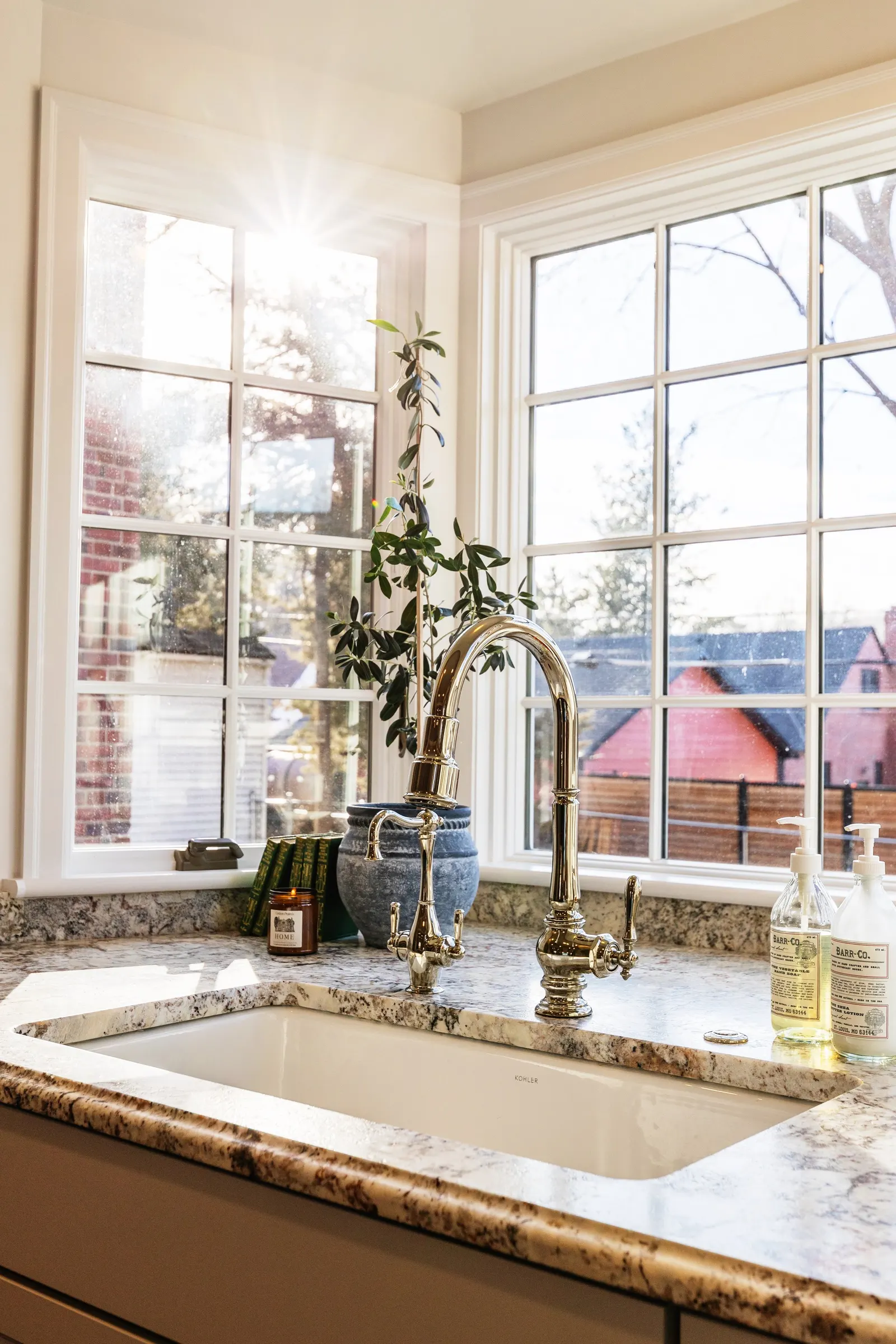 Sunlight coming through a large, corner kitchen window above a marble countertop sink with brass fixtures, decorated with a potted plant, candles, books, and soap dispensers.