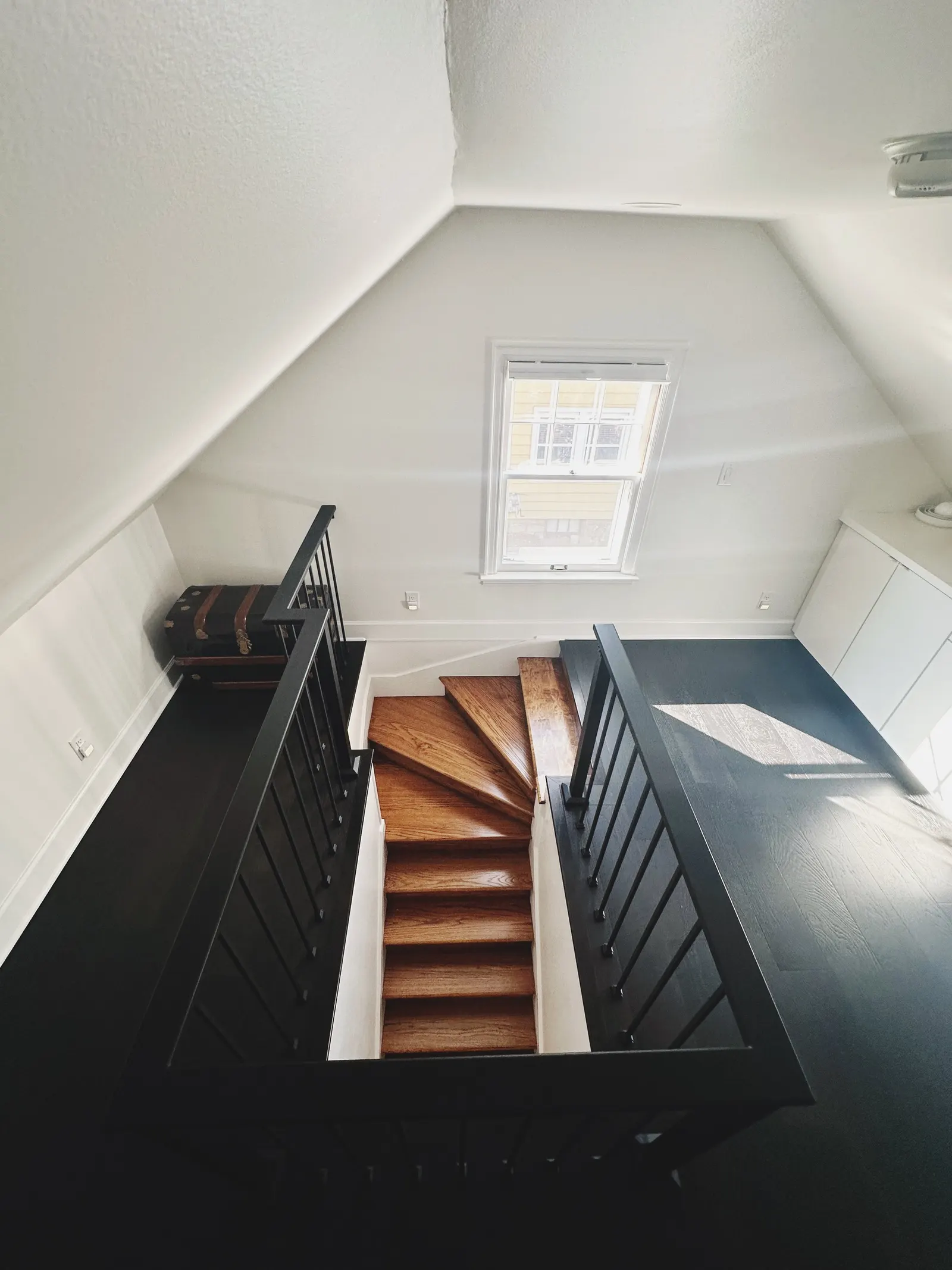 Interior view of a staircase with wooden steps and black railings, leading to an upper floor in a bright, white-walled room with a window allowing sunlight in.