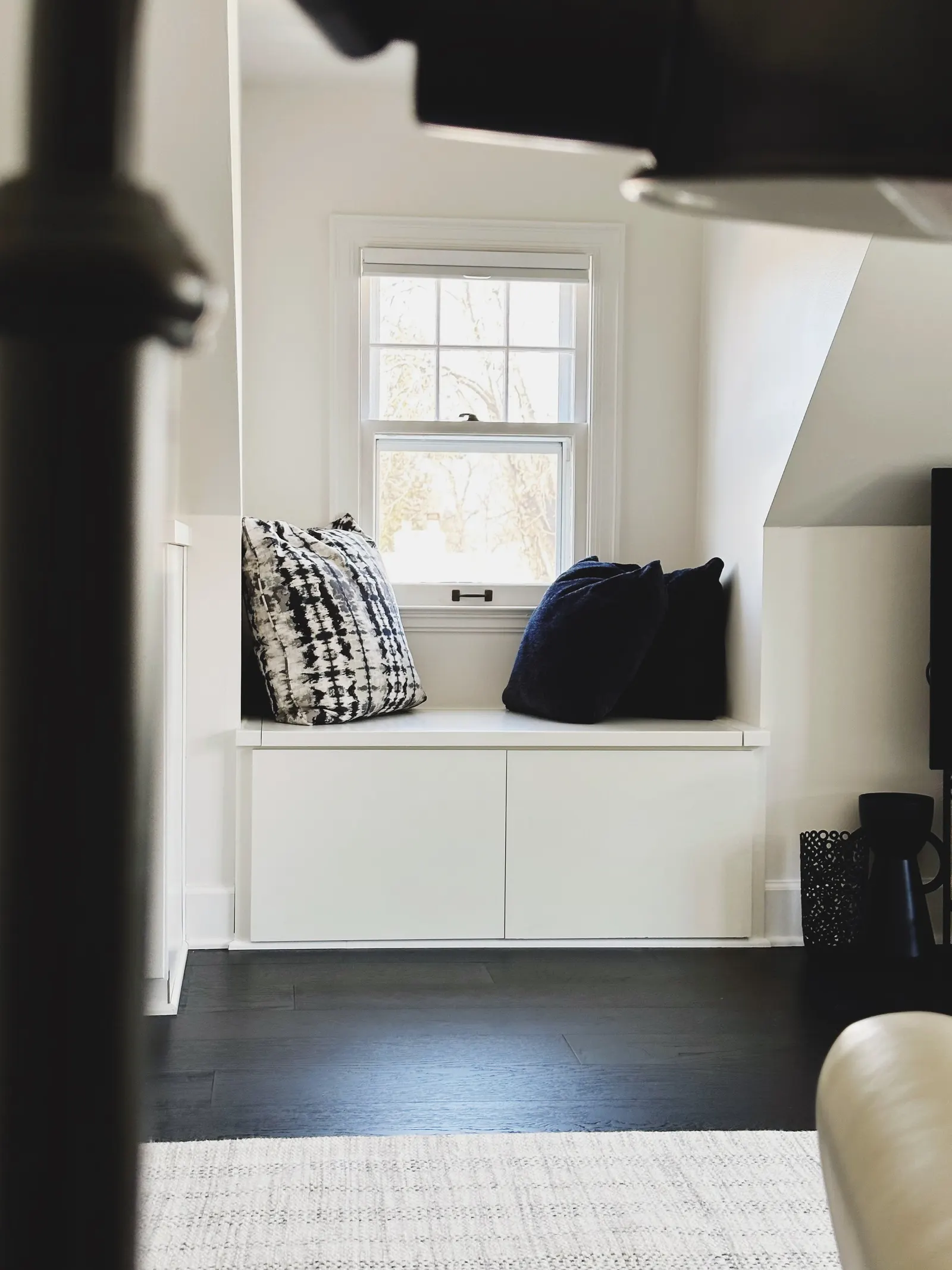 Window seat with three pillows, two dark blue and one patterned black and white, in a bright room with white walls and window, dark hardwood floor, and a beige rug.