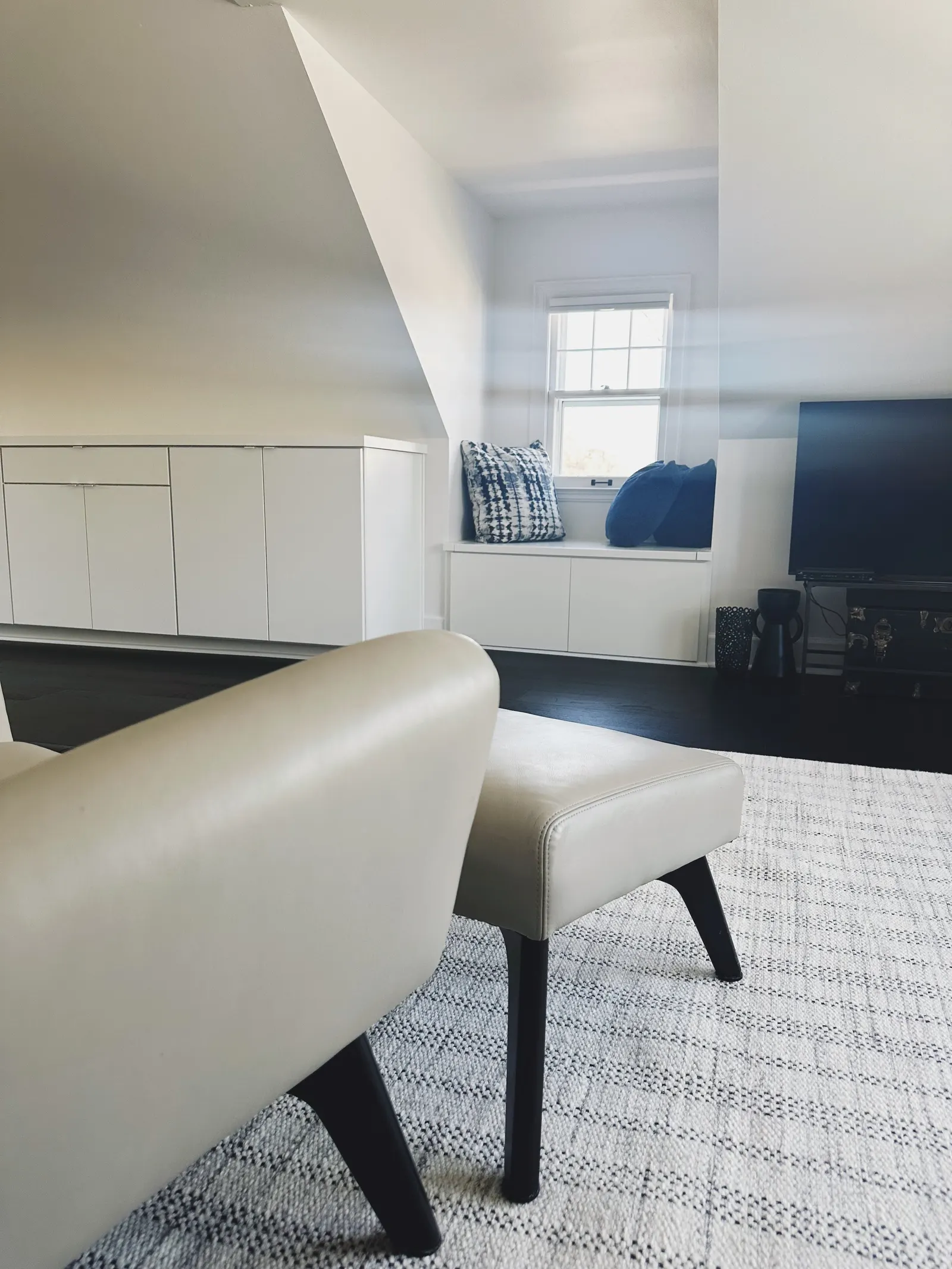 Living room with a white chair and ottoman, a window with cushions, a TV, and a white built-in cabinet under the sloped ceiling.