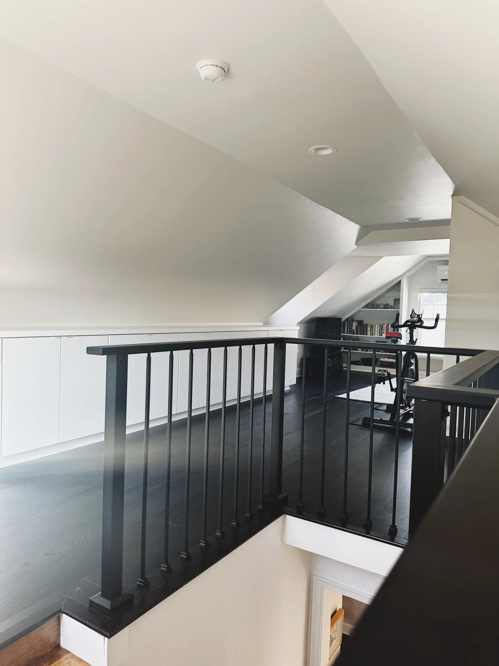 Interior view of a modern upstairs area with black railing, dark wood flooring, white cabinetry, bookshelves, and an exercise bike near a window.