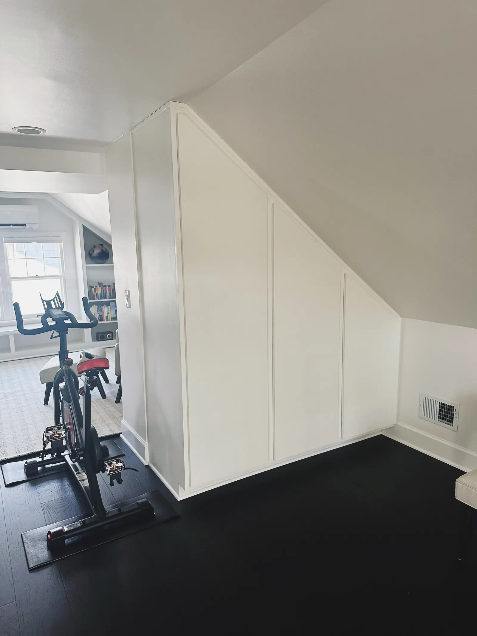 Indoor room with a stationary exercise bike, a window with daylight, a bookshelf with books, and a white wall with decorative molding.