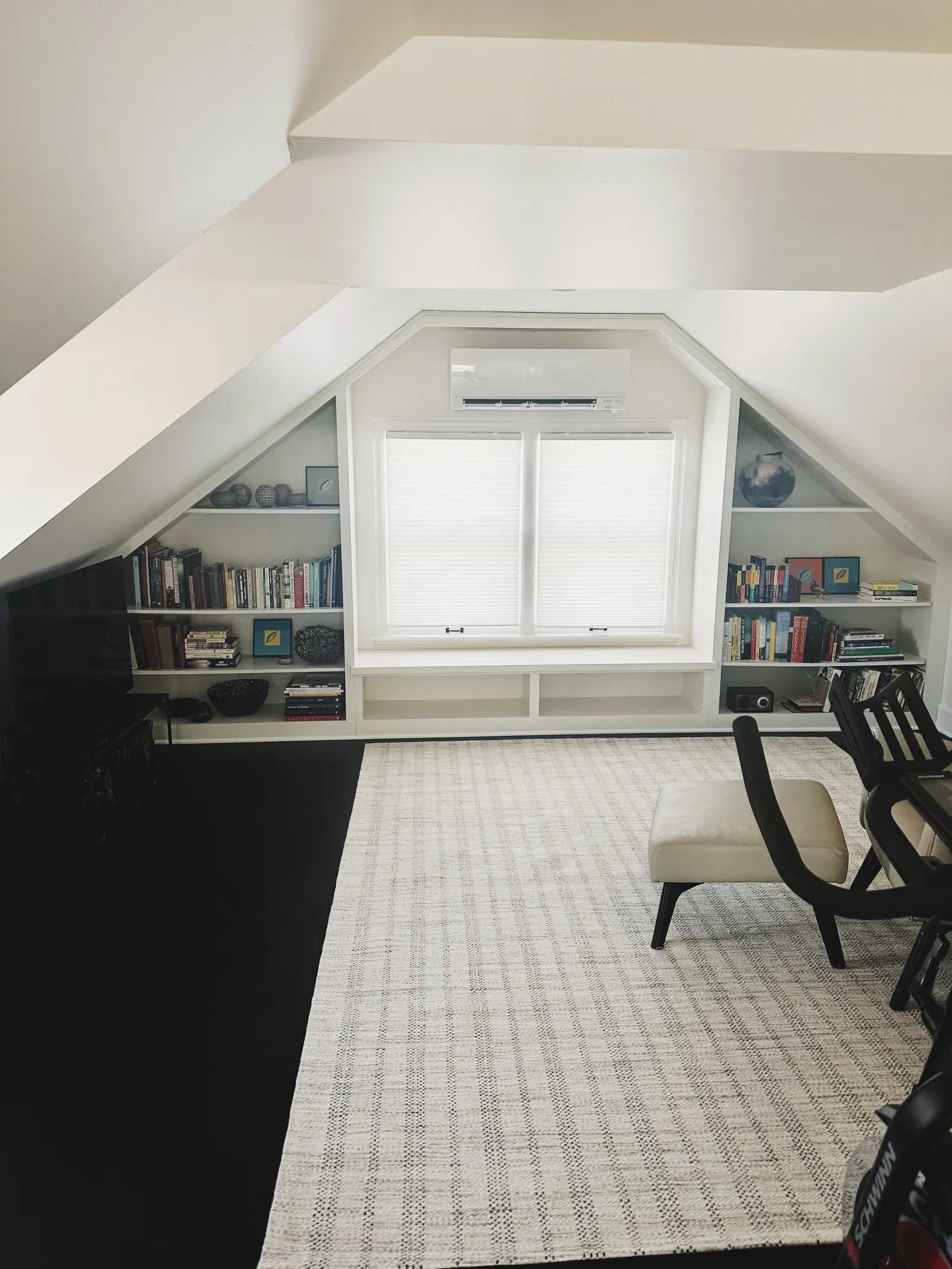 A cozy attic room with built-in white bookshelves on either side of a window, a beige chair, and a black desk in a modern decor setting.