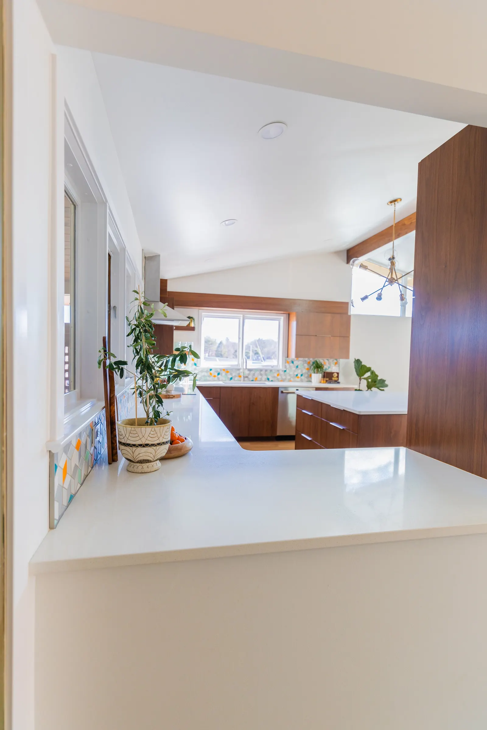 Modern kitchen with wooden cabinets, white countertops, colorful tile backsplash, large window, and a potted plant on the counter.