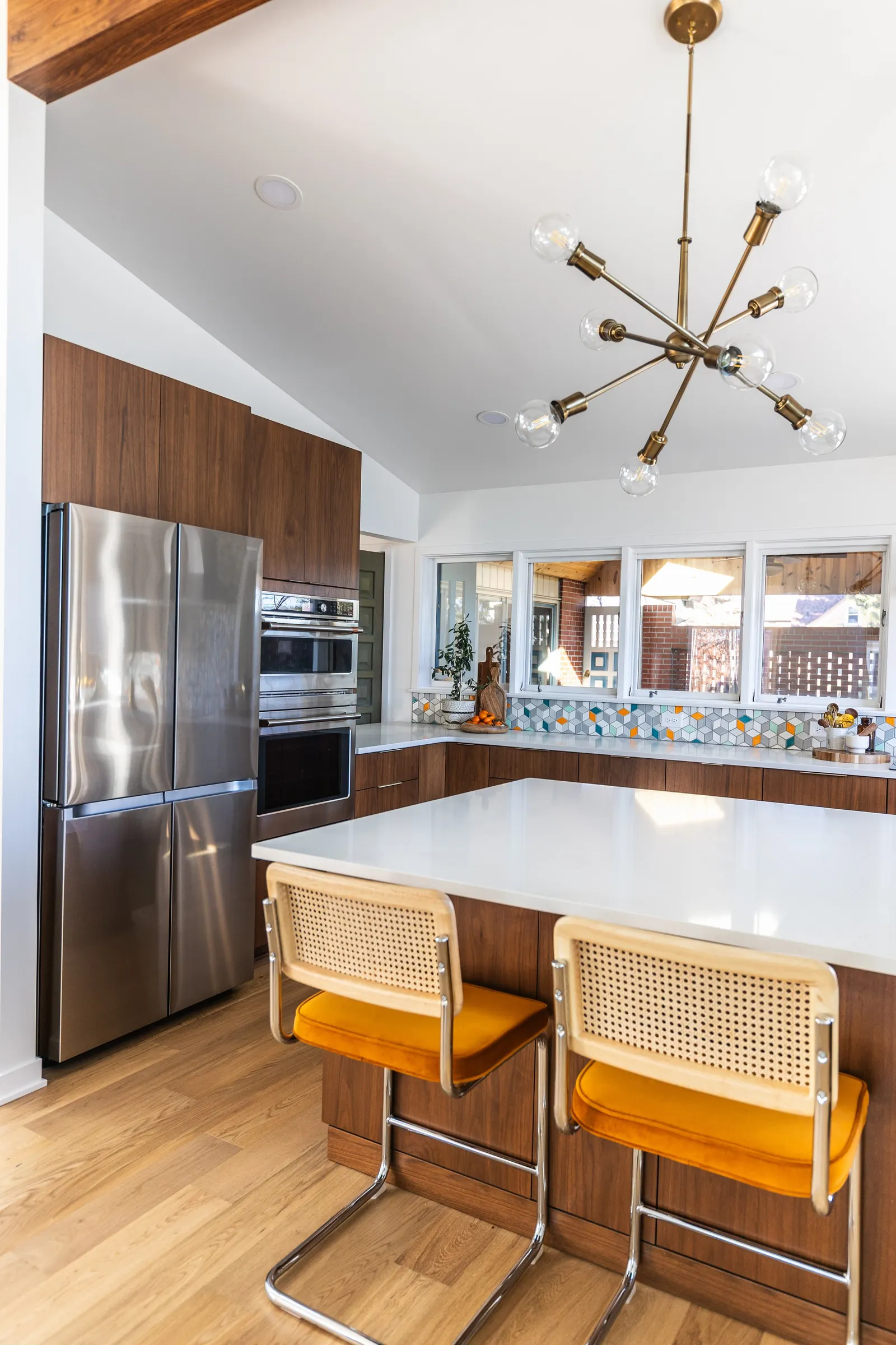 Modern kitchen with a wooden accent wall, stainless steel refrigerator, built-in oven, white countertop, colorful tile backsplash, and two vintage-style bar stools with mustard cushions.