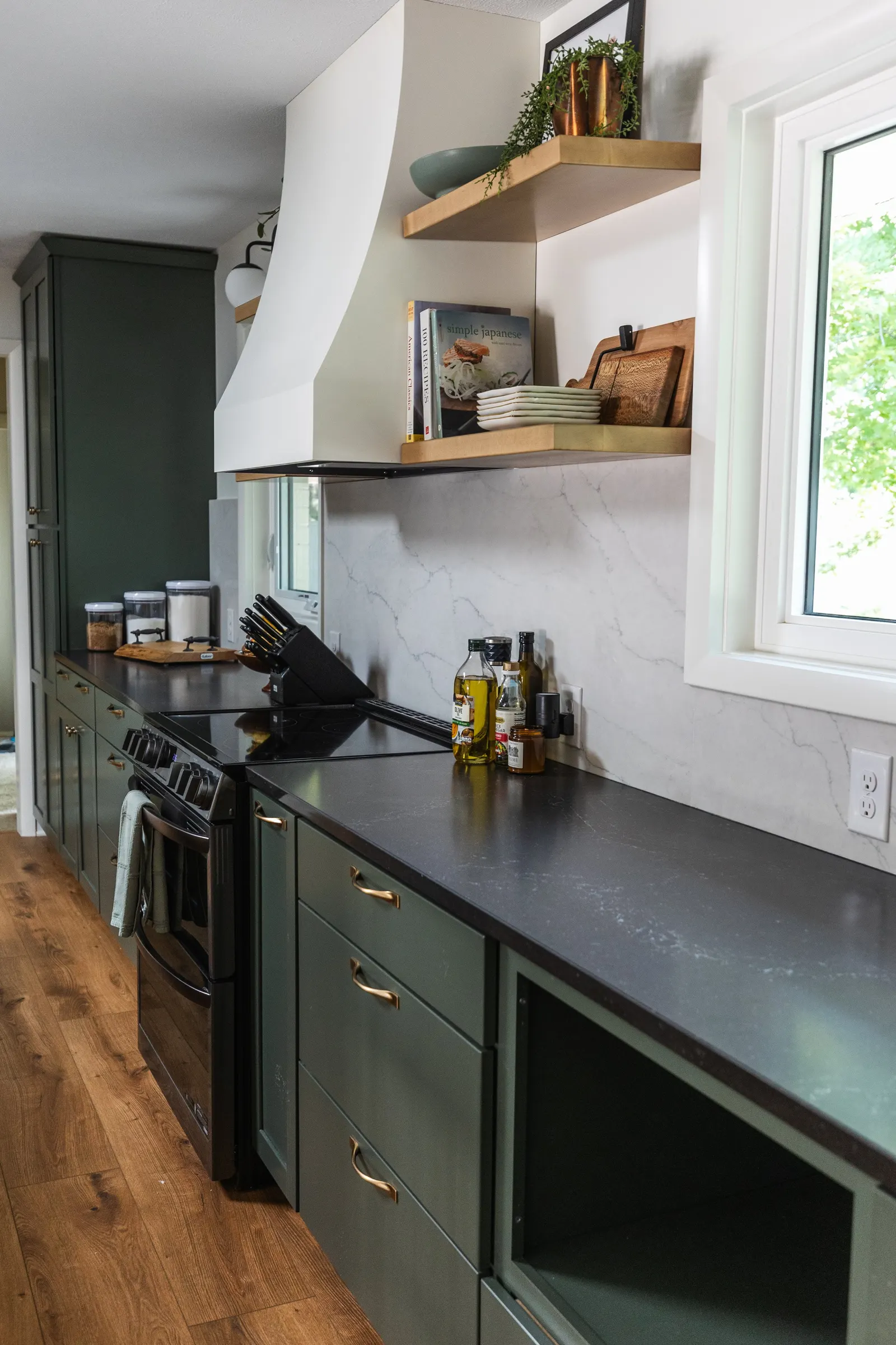 1962 Kitchen Remodel, dark green cabinets, white quartz island, wooden floors, brass hardware, wood cabinets, custom hood, floating shelves, dark countertops, open shelving, large windows, craftsmanship.