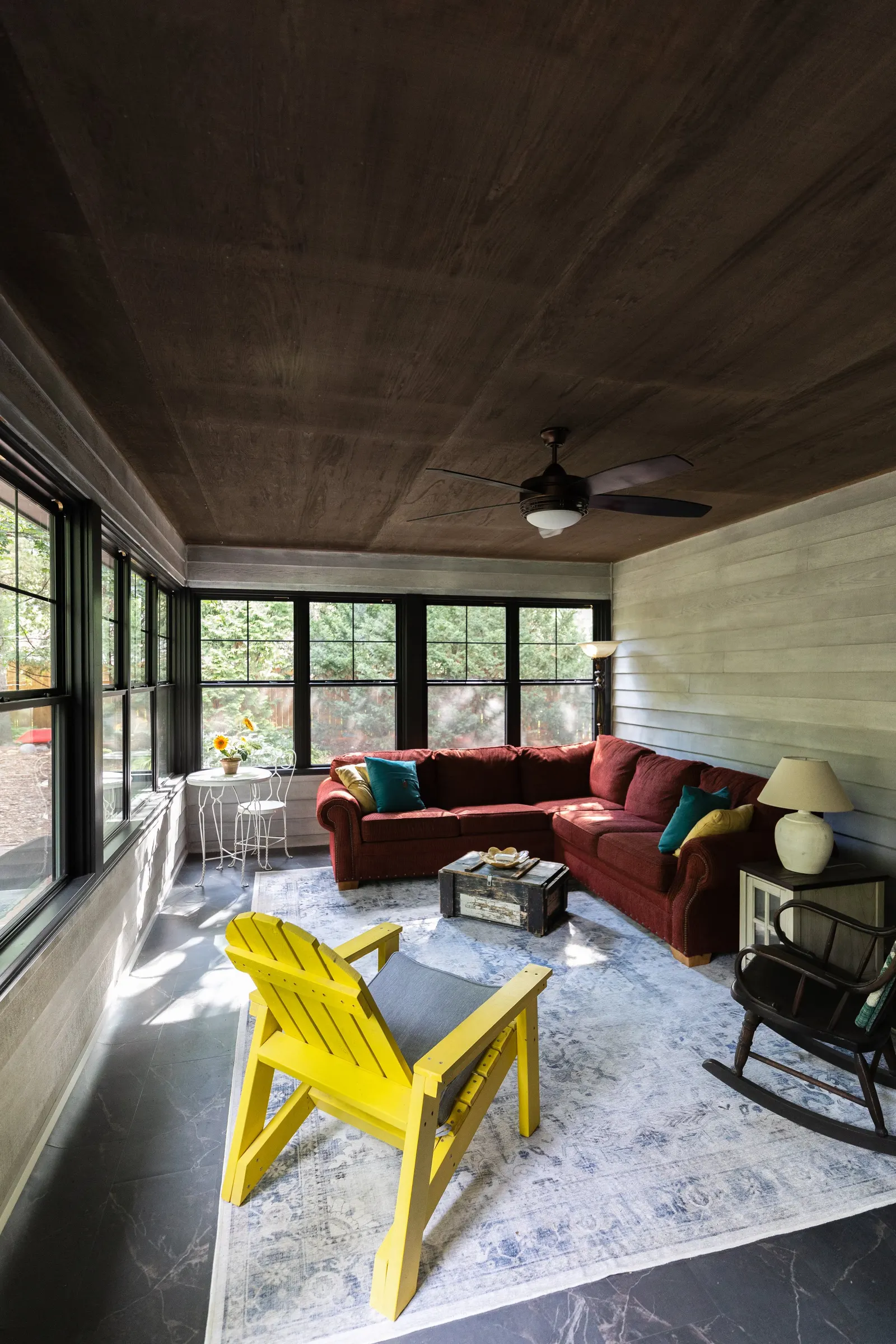 Sunlit living room with a red sectional sofa, yellow Adirondack chair, rocking chair, glass-top table, and a vintage trunk coffee table, surrounded by large windows with black frames and a ceiling fan.
