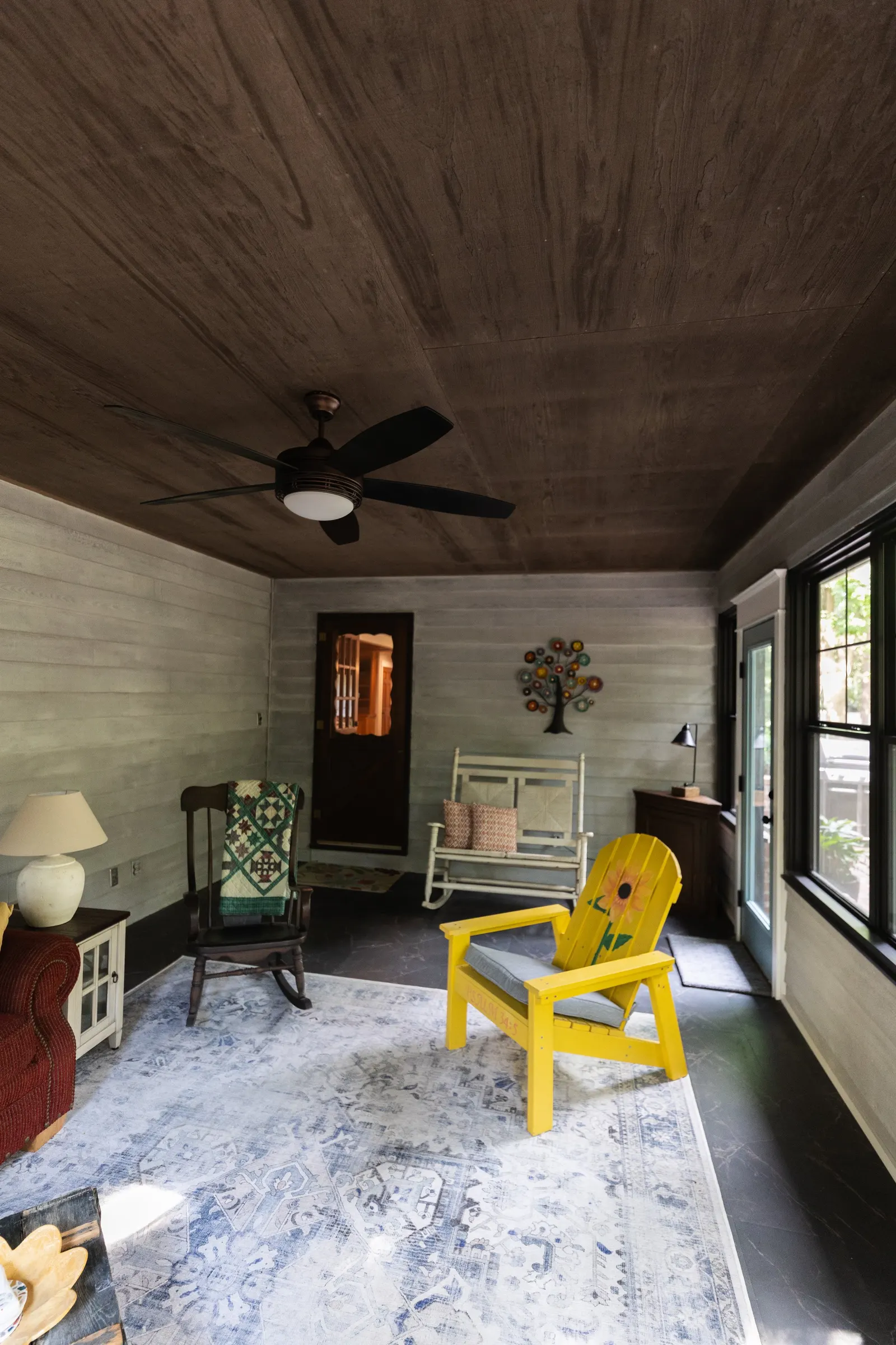 Living room with light gray wood-paneled walls, dark wood ceiling with a black ceiling fan, large windows, and a mix of chairs including a yellow Adirondack chair with a sunflower painted on it, and other chairs with patterned cushions. There's a whi