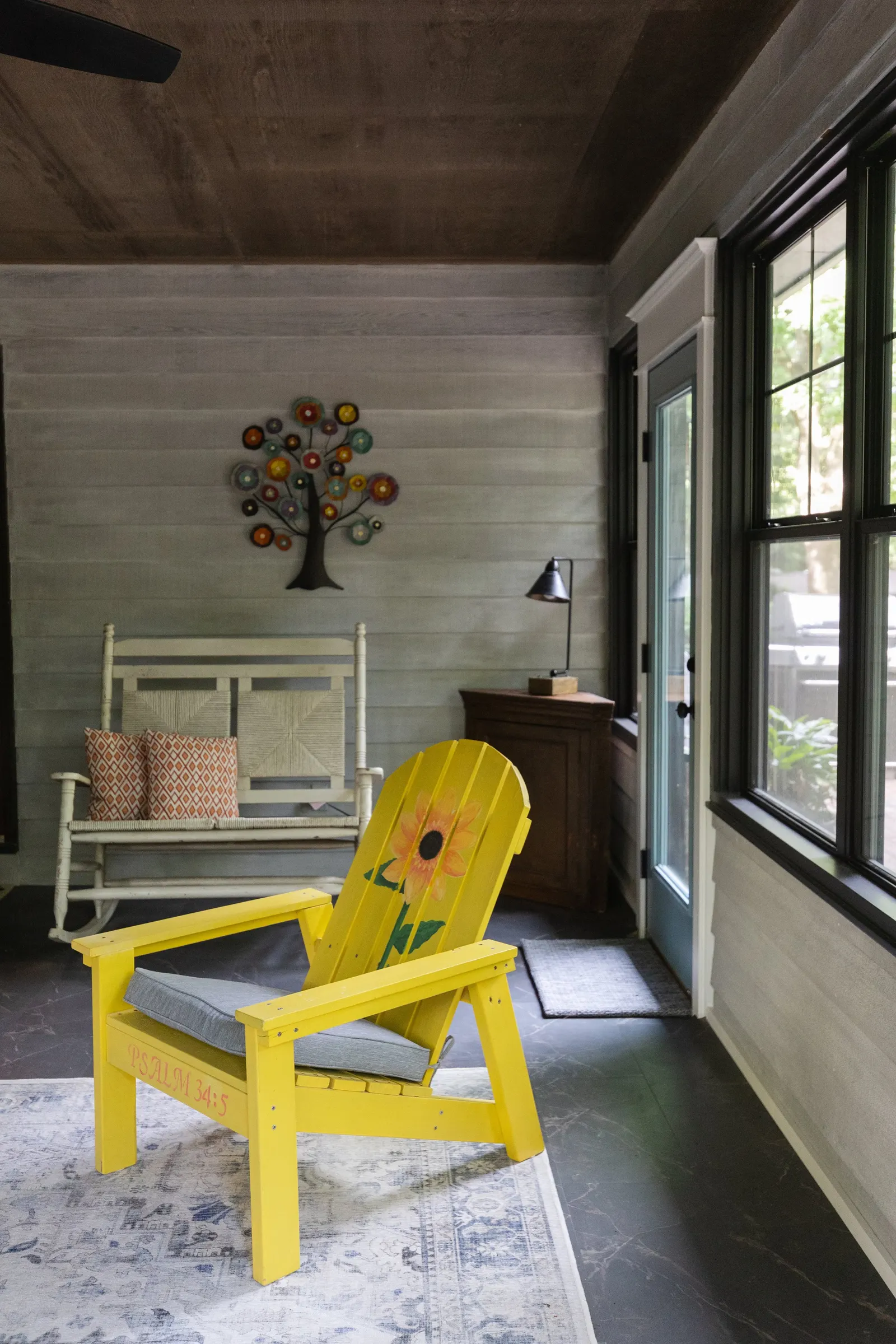 Sunroom with a yellow Adirondack chair, a white wooden bench with orange patterned pillows, a wall decoration of a colorful tree, a wooden cabinet with a black desk lamp, and large windows with black frames.