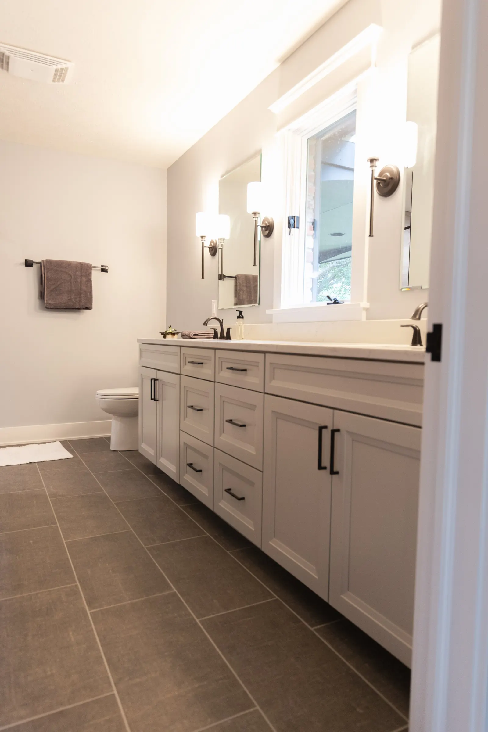 Modern bathroom showcasing a double sink vanity with light-colored cabinetry and black handles, a large mirror, wall-mounted light fixtures, a window above the vanity, a towel hanging on a bar, and a toilet against a white wall.