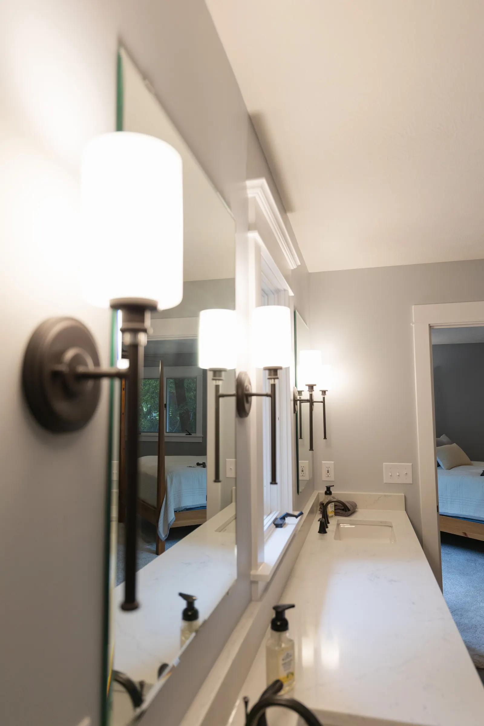 Bathroom vanity with a large mirror, two wall-mounted light fixtures, and bathroom items on the counter, with a bedroom visible through the doorway.