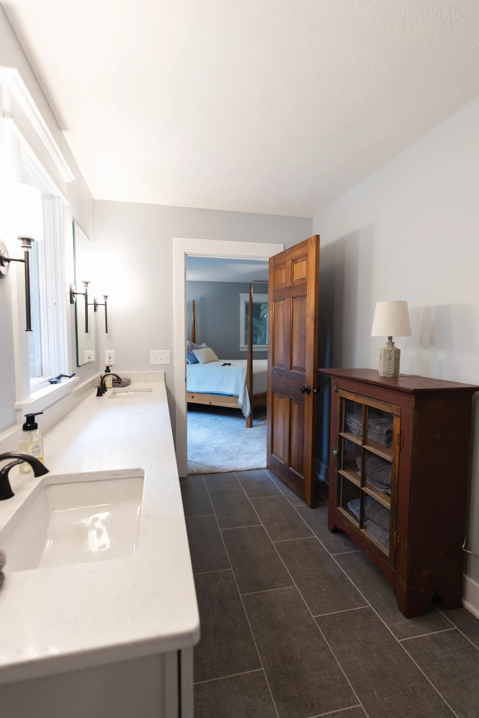 Bathroom with two sinks, a mirror, a wooden cabinet with glass doors, and an open wooden door leading to a bedroom with a four-poster bed.