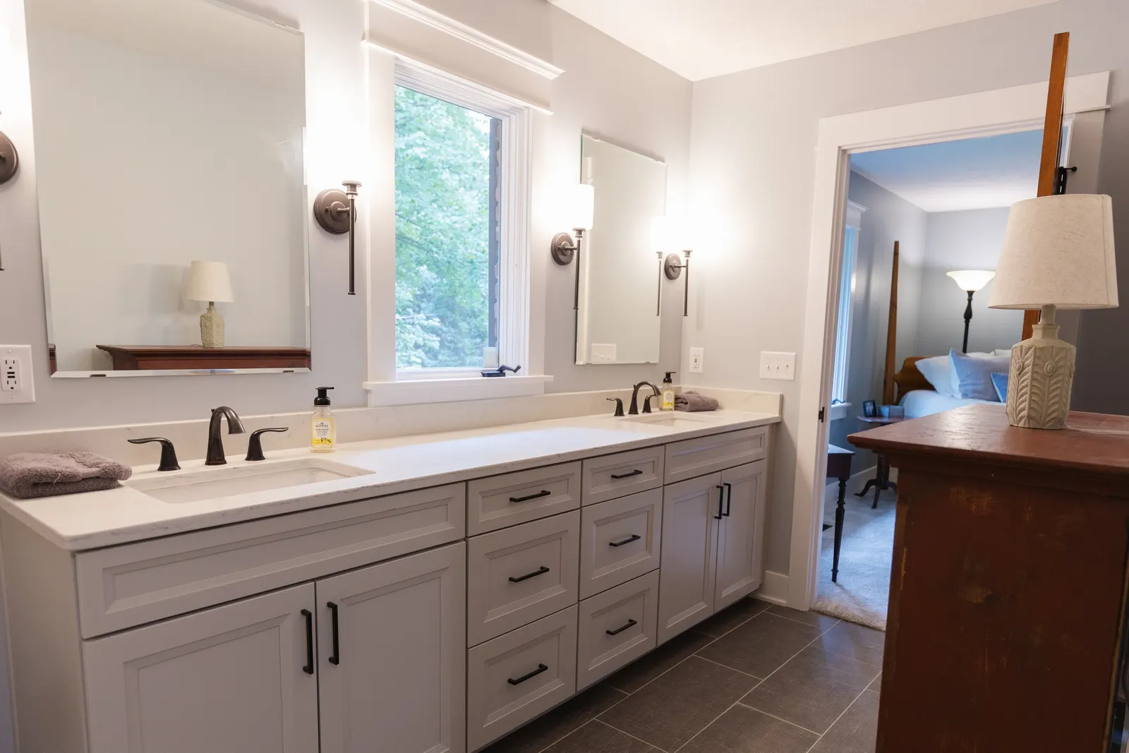 Bathroom with double vanity, large mirror, wall-mounted lights, window showing greenery, and doorway to bedroom with bed and bedside table