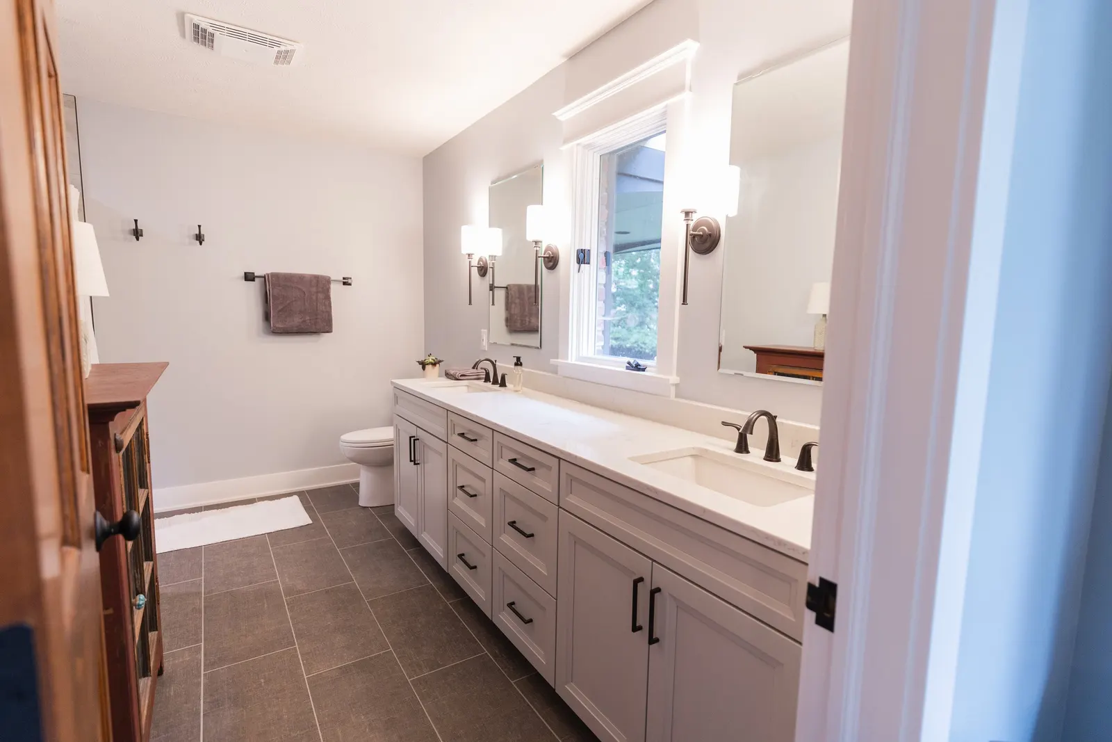 Modern bathroom with double vanity, white cabinets, marble countertop, two sinks, wall-mounted light fixtures, mirror, window, toilet, and brown towels.