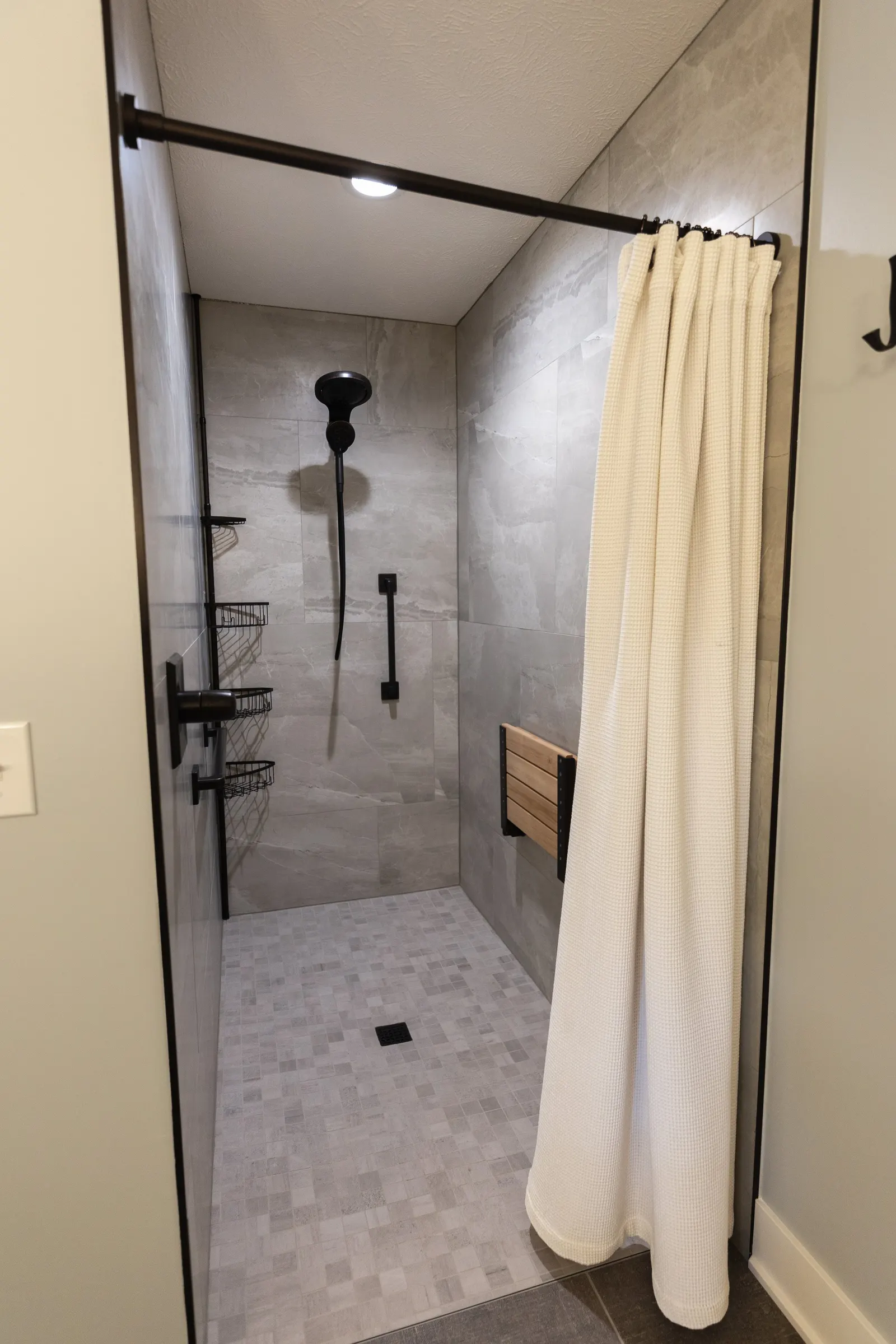 Modern walk-in shower with gray tiled walls and floor, black fixtures, a beige curtain on a metal rod, and a wooden fold-down seat.