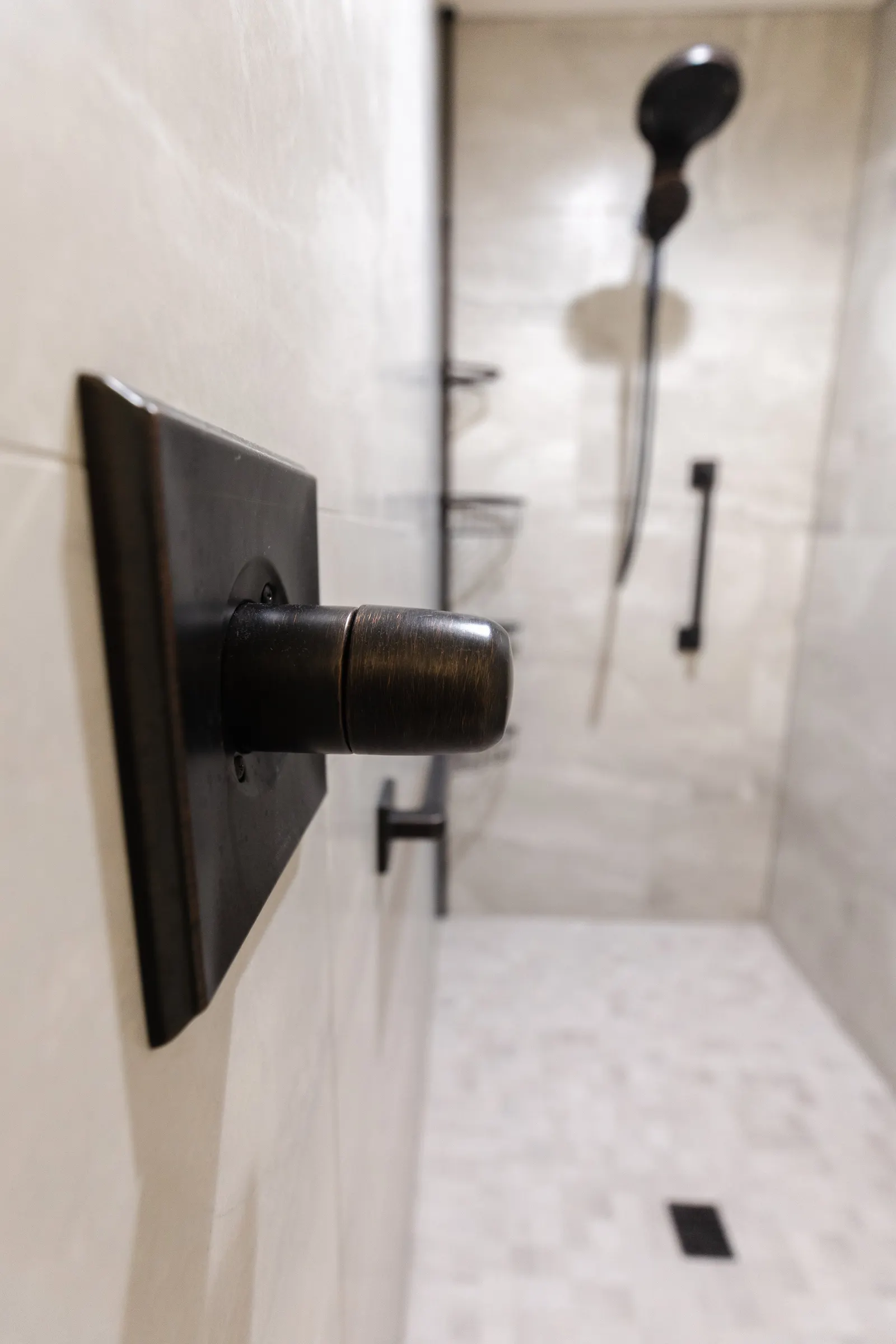 Close-up of a bronze shower handle with a walk-in shower in the background, featuring a black showerhead, a handheld shower wand, and shelving in a tiled bathroom.