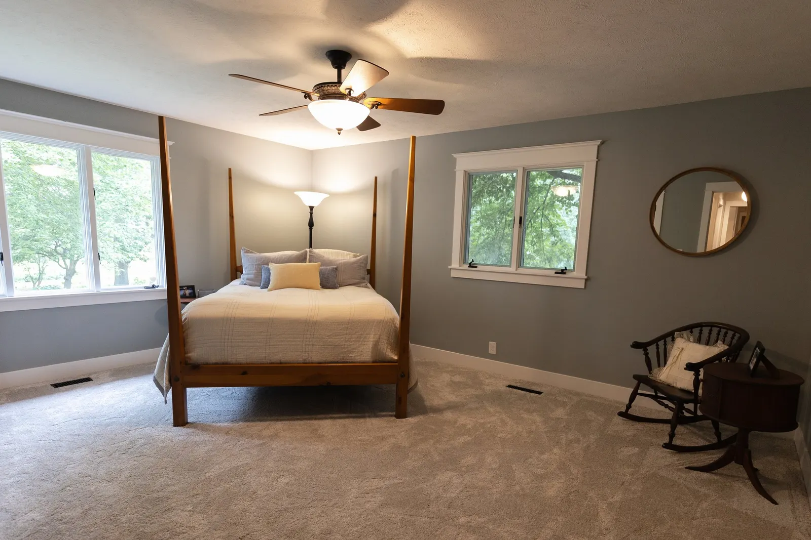 A bedroom with light gray walls, beige carpet, a four-poster wooden bed with neutral bedding, a window with trees outside, a black floor lamp behind the bed, a small wooden rocking chair with a pillow, and a round wall mirror.