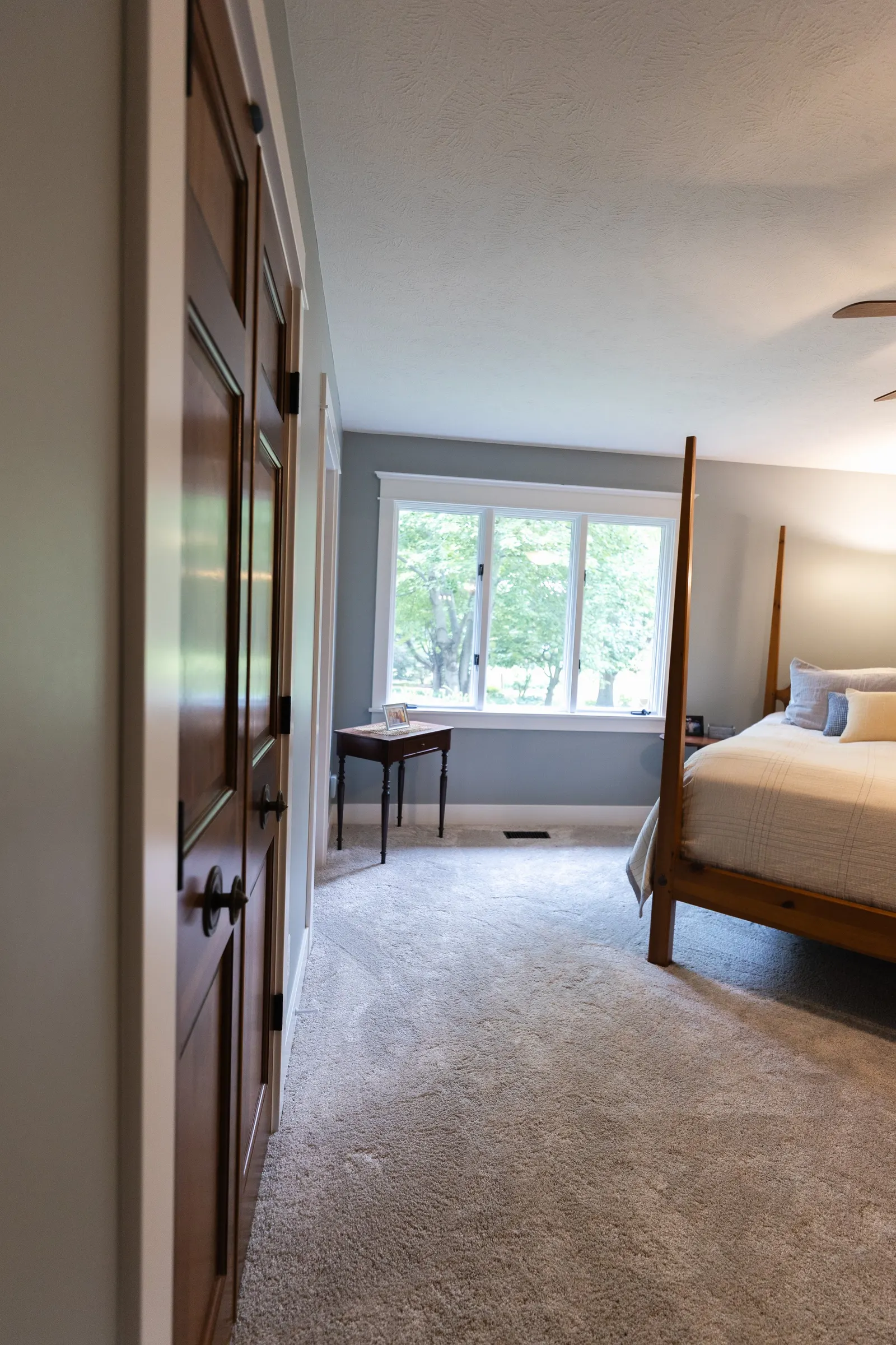 A bedroom with a large window, a wooden bed frame, a small table with a photo, and beige carpet.