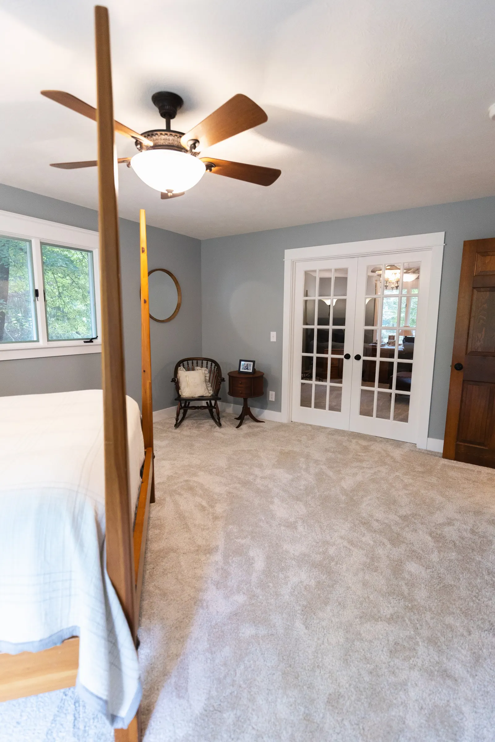 A bedroom with a bed on the left side, a window with green trees outside, a ceiling fan with lights, a small side table, and a chandelier visible through glass double doors.