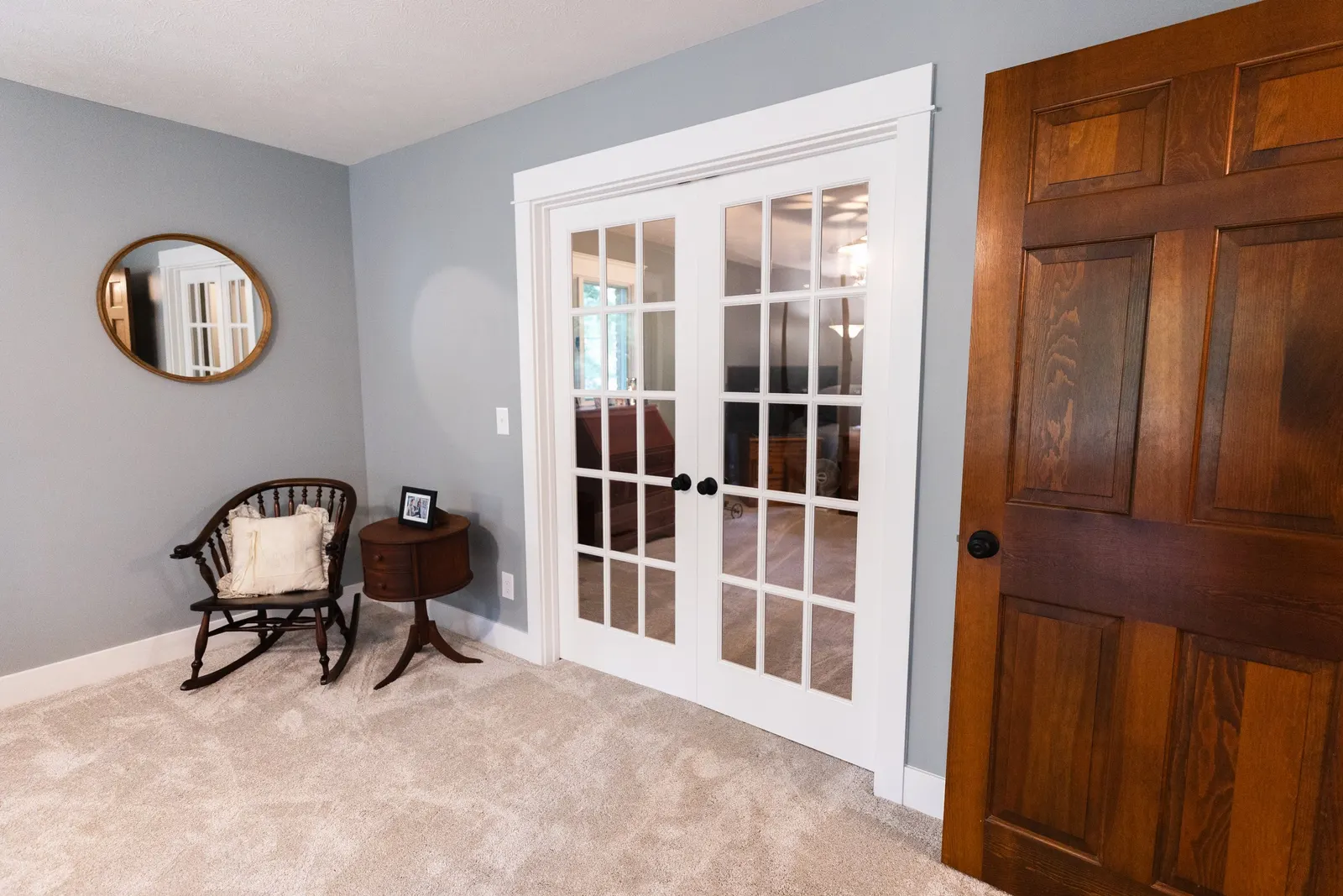 Living room with light gray walls, beige carpet, a round mirror, a wooden chair with a pillow, a small wooden side table with a framed photo, white French doors, and a wooden door.