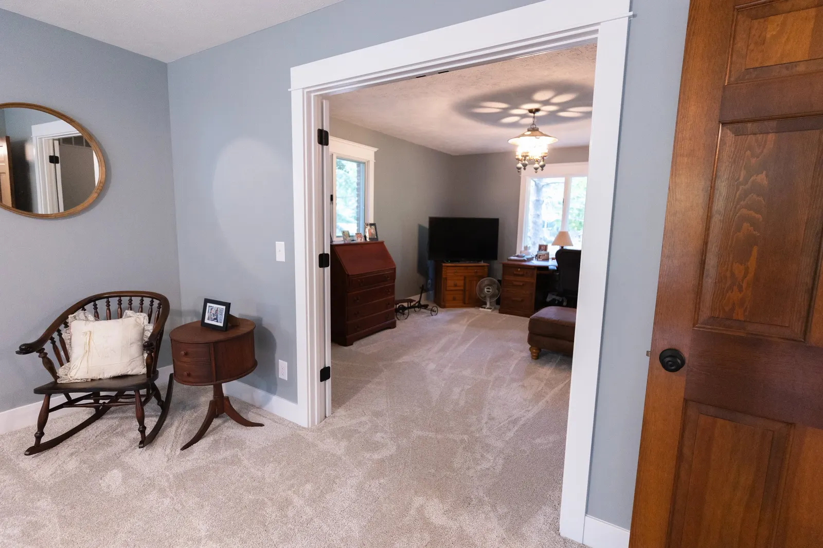 View of a living room with open door, beige carpet, wooden furniture, blue walls, and a chair with pillows outside the doorway.