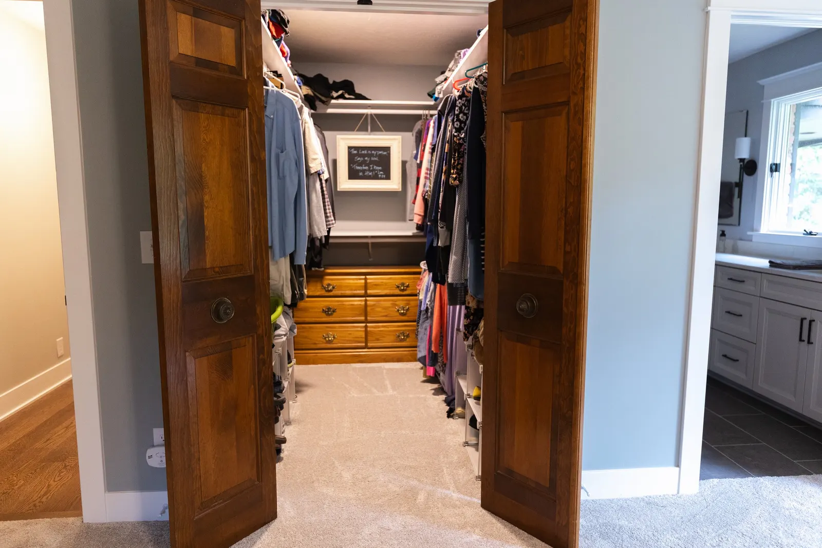 View into a walk-in closet with double doors open, containing hanging clothes, a chest of drawers at the back, and shelves on both sides. Adjacent to the closet is a partial view of a room with a vanity and a window.