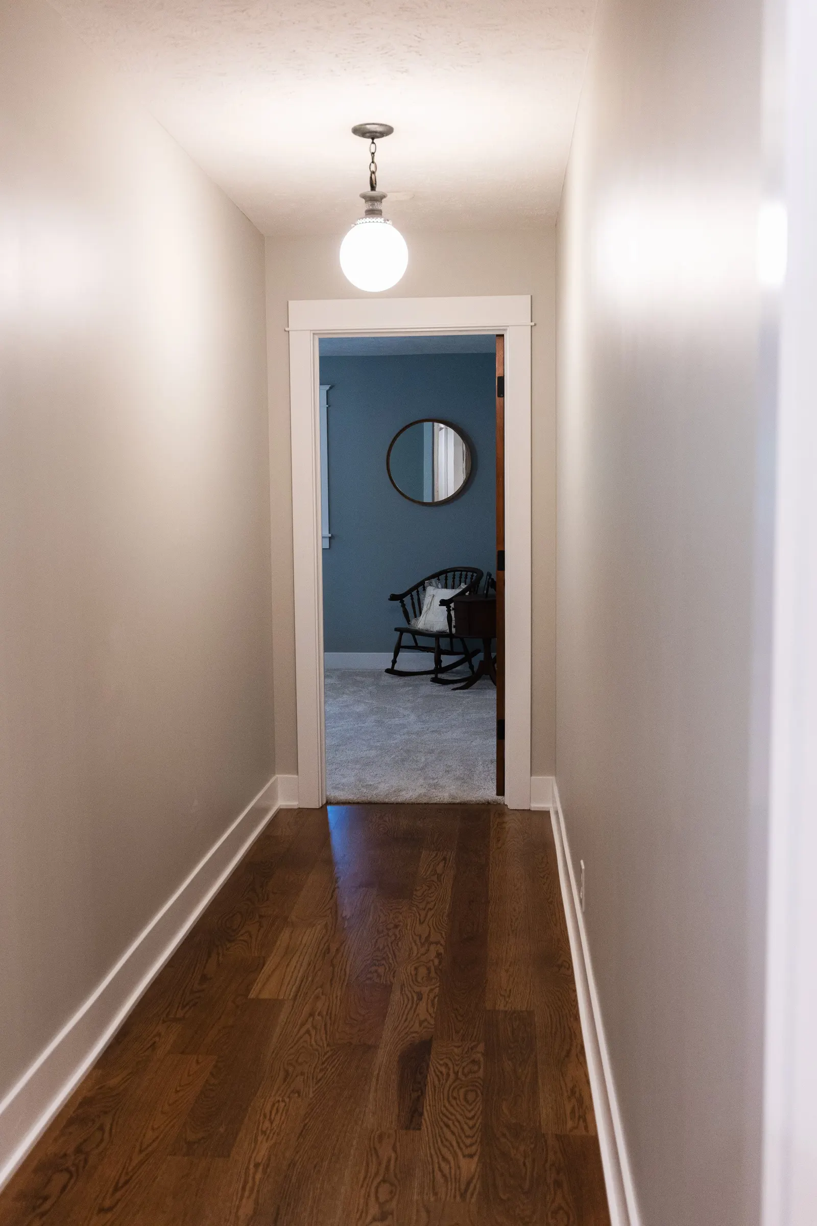 Empty narrow hallway with beige walls, dark hardwood floor, white baseboards, and a ceiling light fixture leading into a room with a blue wall, a black rocking chair, a mirror, and a window.