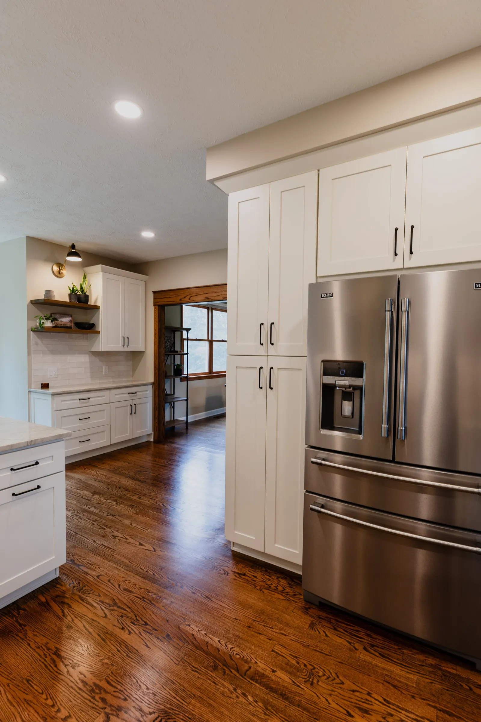 Modern kitchen with stainless steel refrigerator, white cabinets, wooden floor, and a sitting area with window.