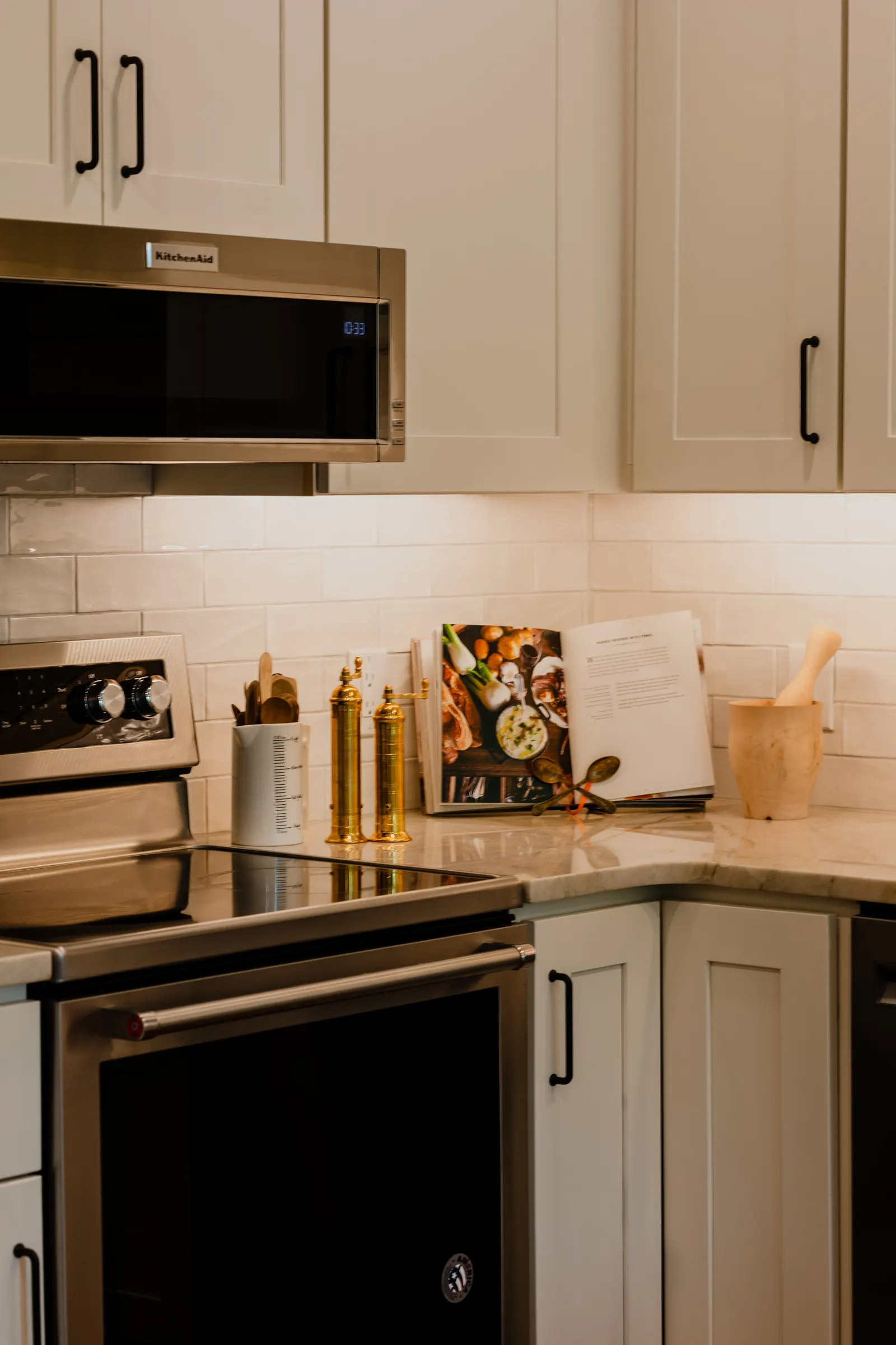 A modern kitchen corner with white cabinets, a stainless steel microwave, a stove with a marble countertop, cookbooks, and kitchen utensils.