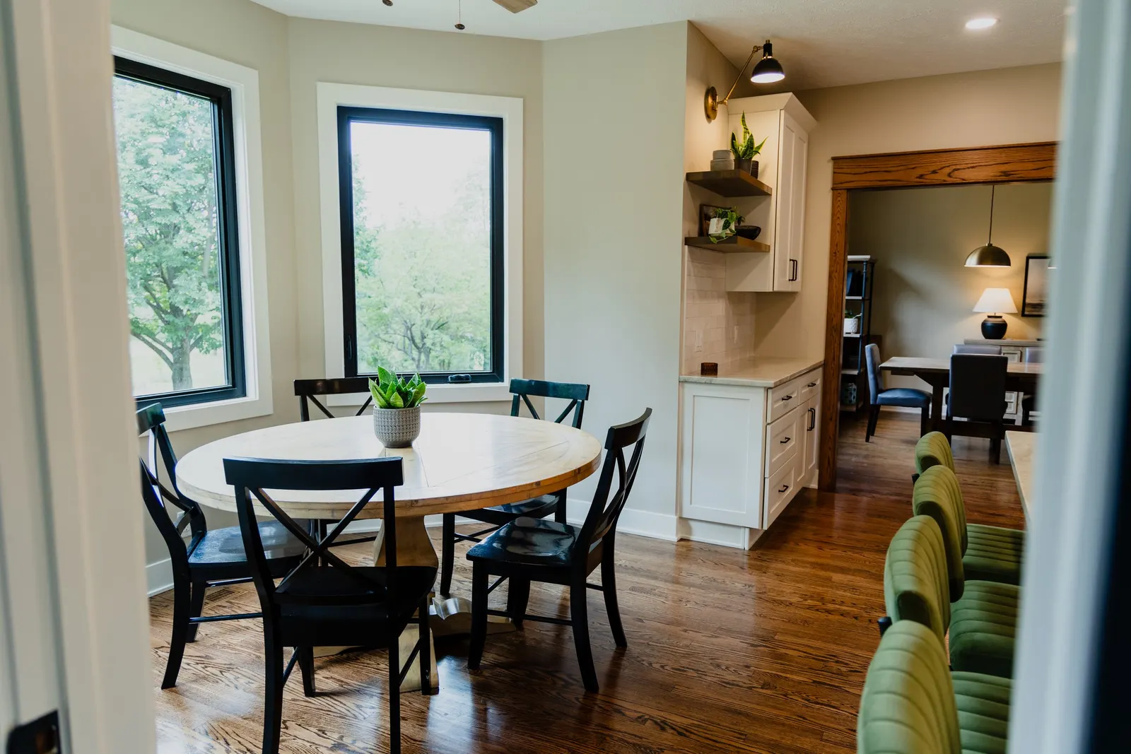 A dining area with a round wooden table, black chairs, green bar stools, large windows with view of trees, and a kitchen in the background with white cabinets and open shelving.