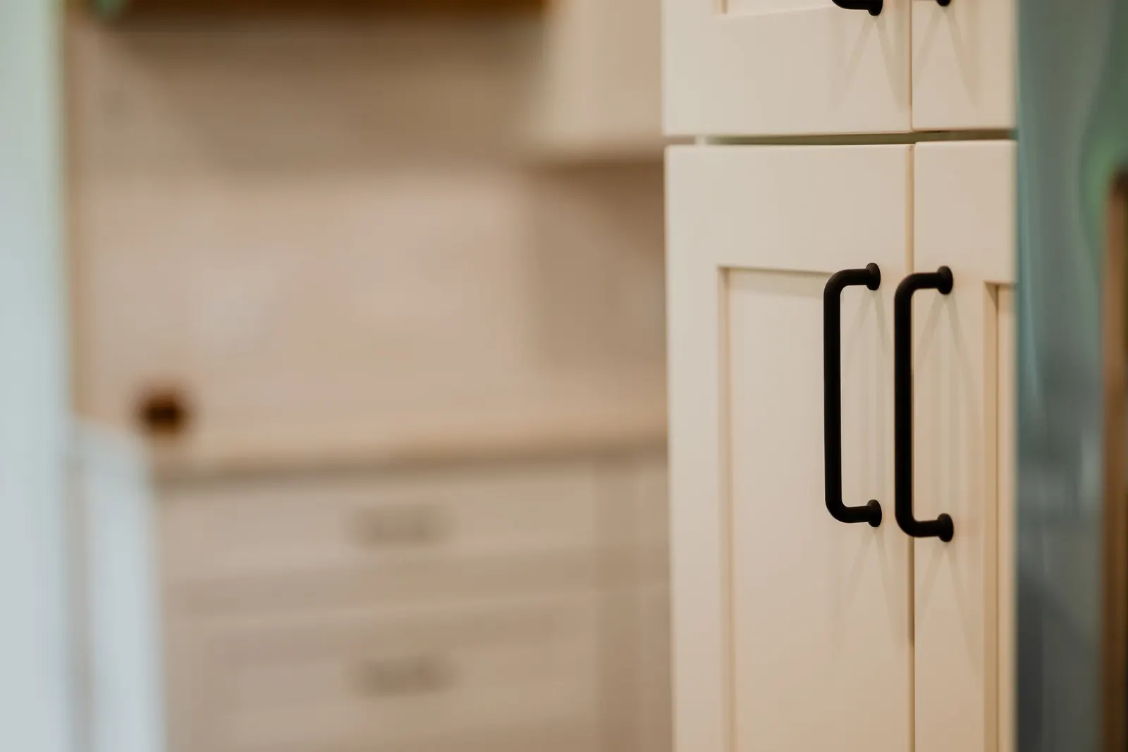 Close-up of white kitchen cabinet doors with black handles, blurred surrounding cabinets. 1935 Kitchen Remodel. Black Matte Hardware. White Cabinets.