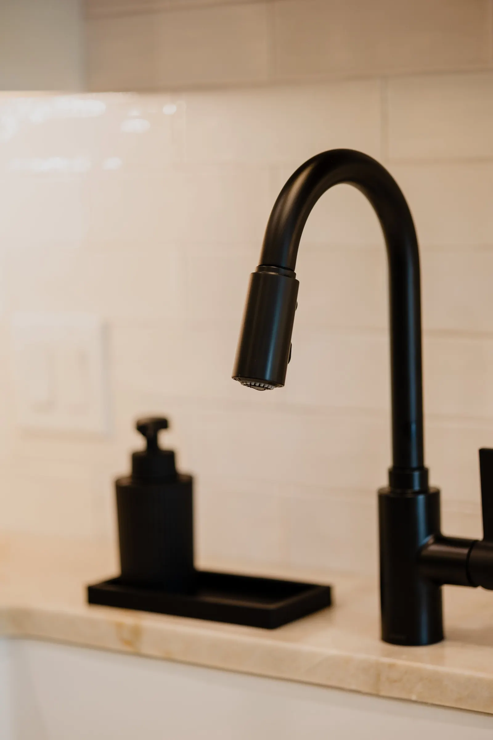 Close-up of a black kitchen faucet and a black soap dispenser on a beige countertop against a white tile wall. 1935 Kitchen Black Matte Faucet. White Subway Tile.