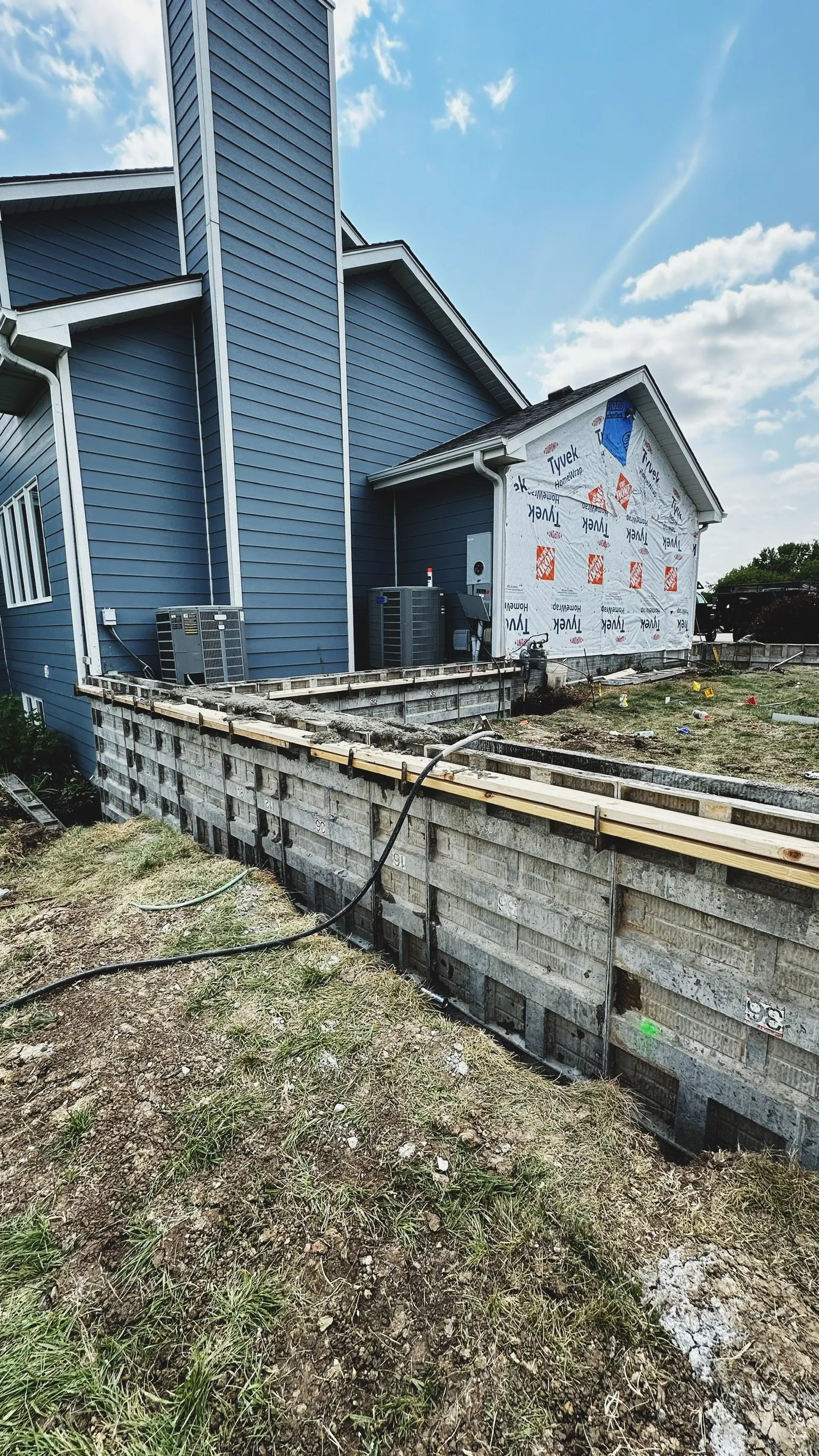 Rear of a blue house under construction with a concrete foundation, construction materials, and equipment visible, and a partly cloudy sky overhead.