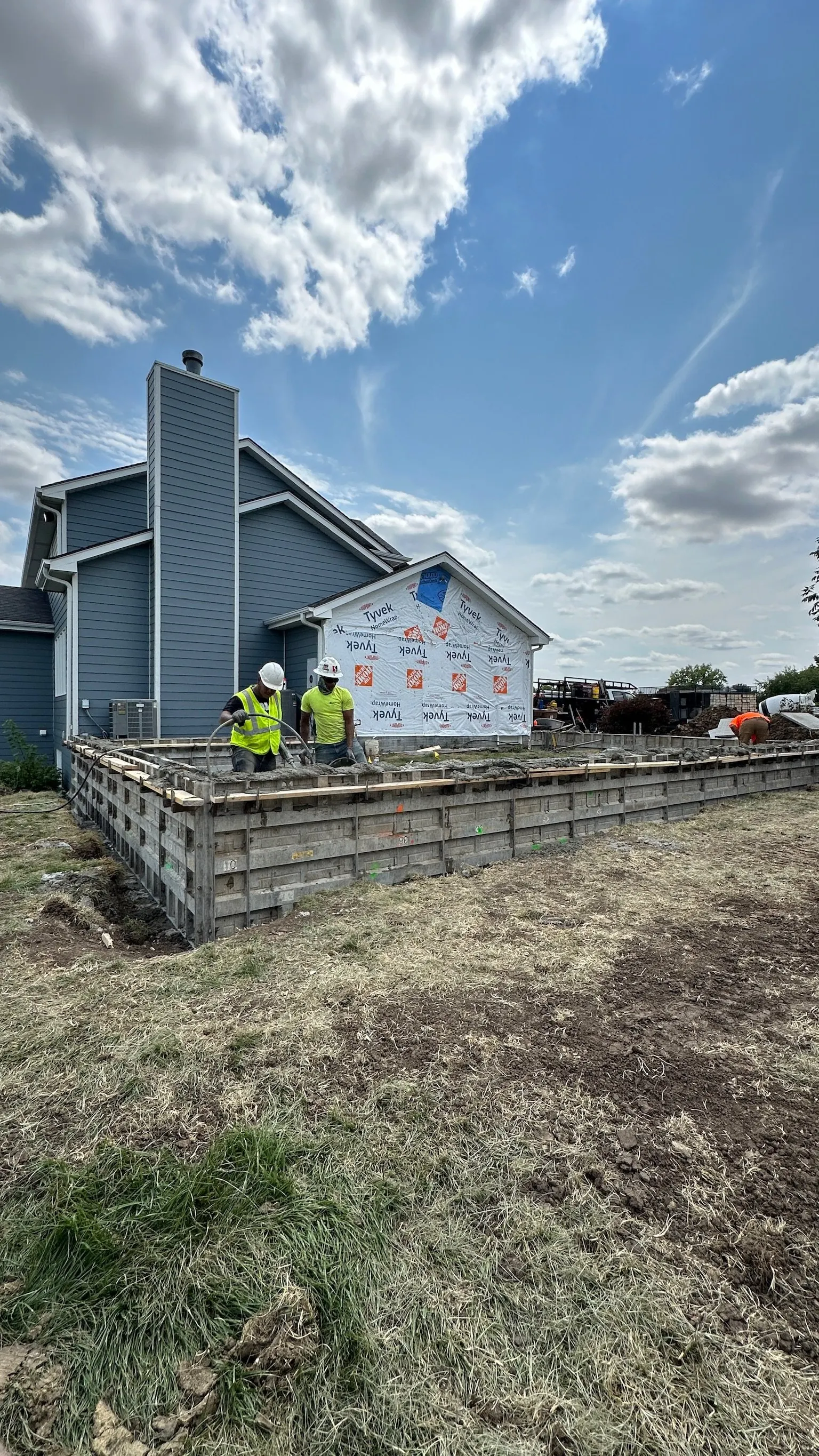 Construction workers pouring concrete on a house foundation under a partly cloudy sky.