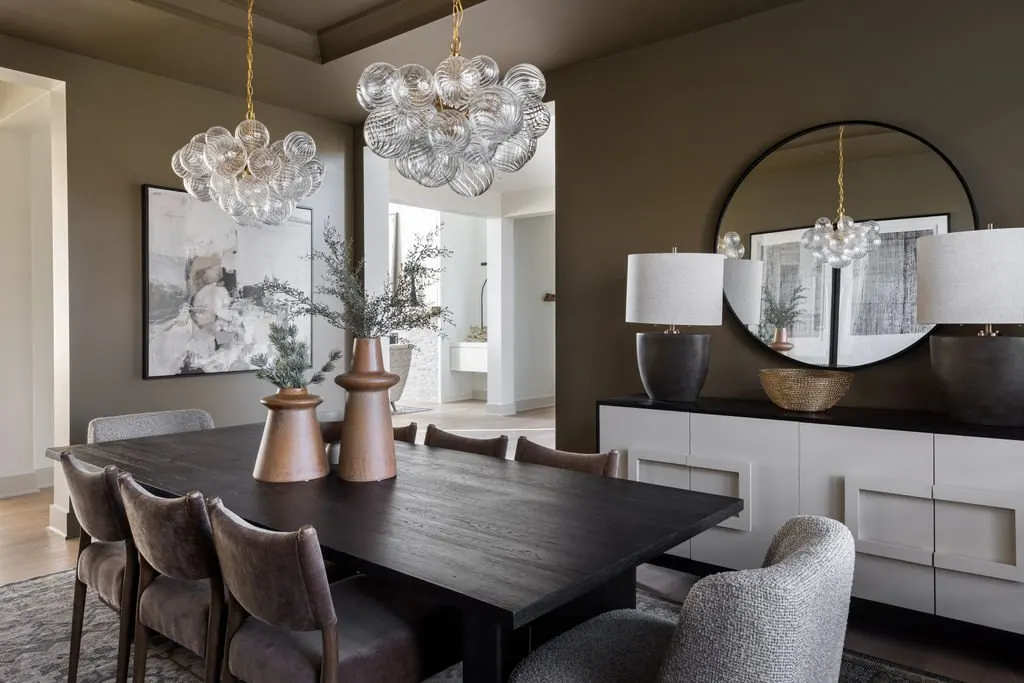 Modern dining room with a black wooden table, eight upholstered chairs, three bronze vases with branches on the table, hanging bubble chandeliers, a large round mirror on the wall, and a white sideboard with two lamps and a basket.