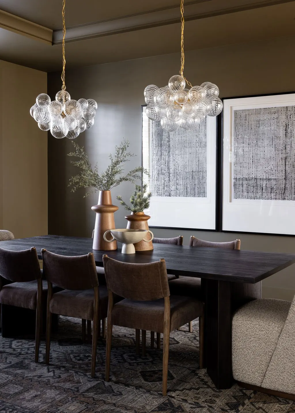 A dining room with a dark wooden table, six brown upholstered chairs, and a beige armchair. Two vases with greenery and a decorative bowl are on the table. Two modern glass bubble pendant lights hang above. Two large framed abstract black and white a