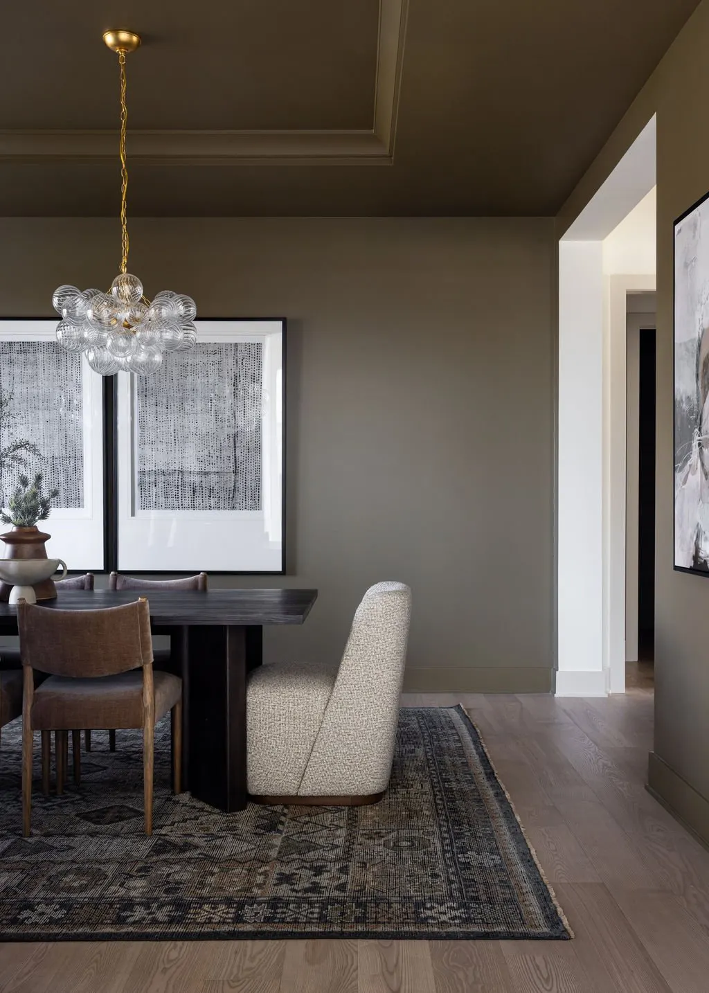 Interior dining room with a dark wooden table, beige upholstered chairs, a decorative rug, a modern glass chandelier, and framed artwork on the walls, with a window to the left.